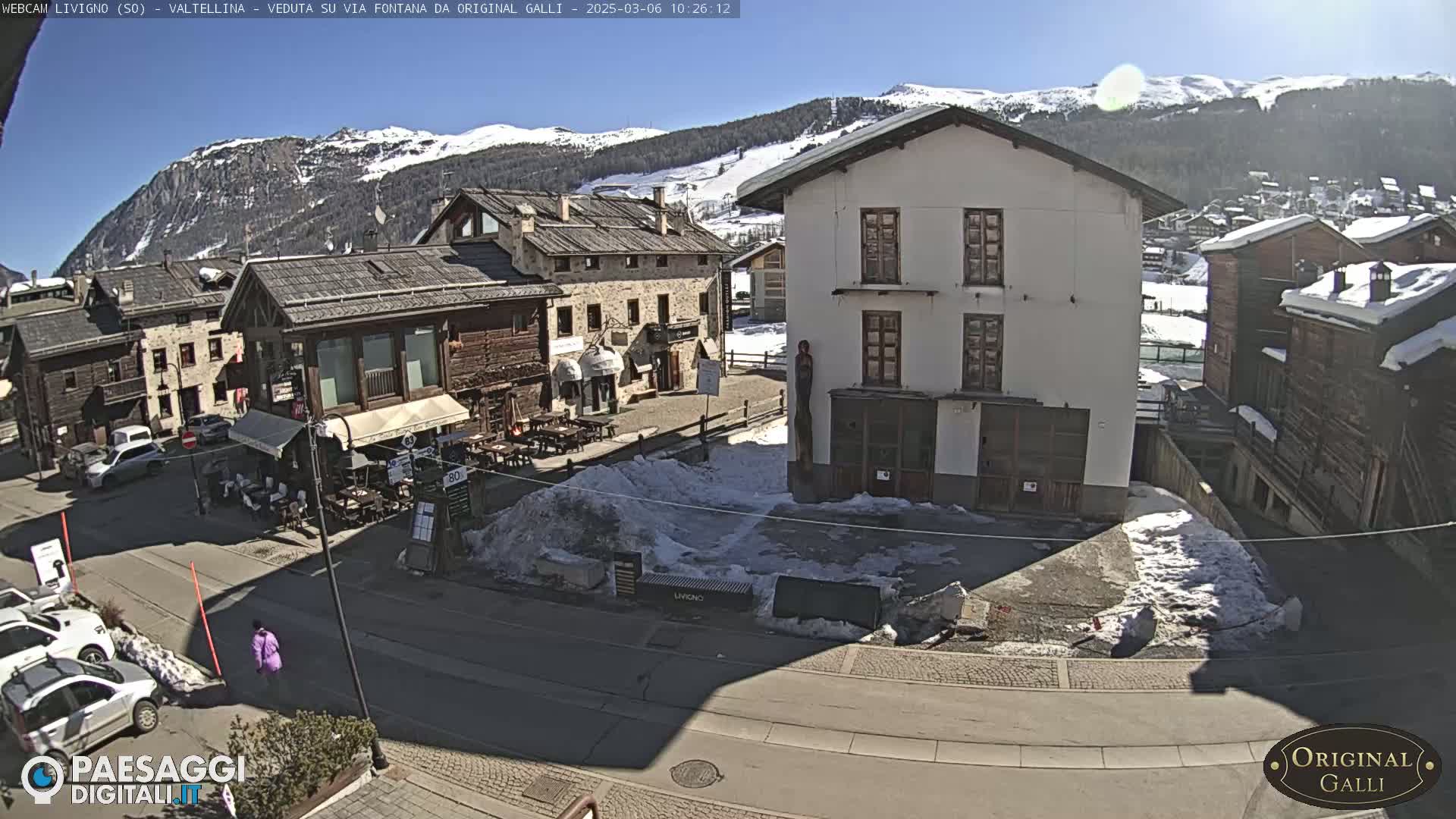 A sunny mountain village square with snow patches, several buildings, and cars parked along the street.