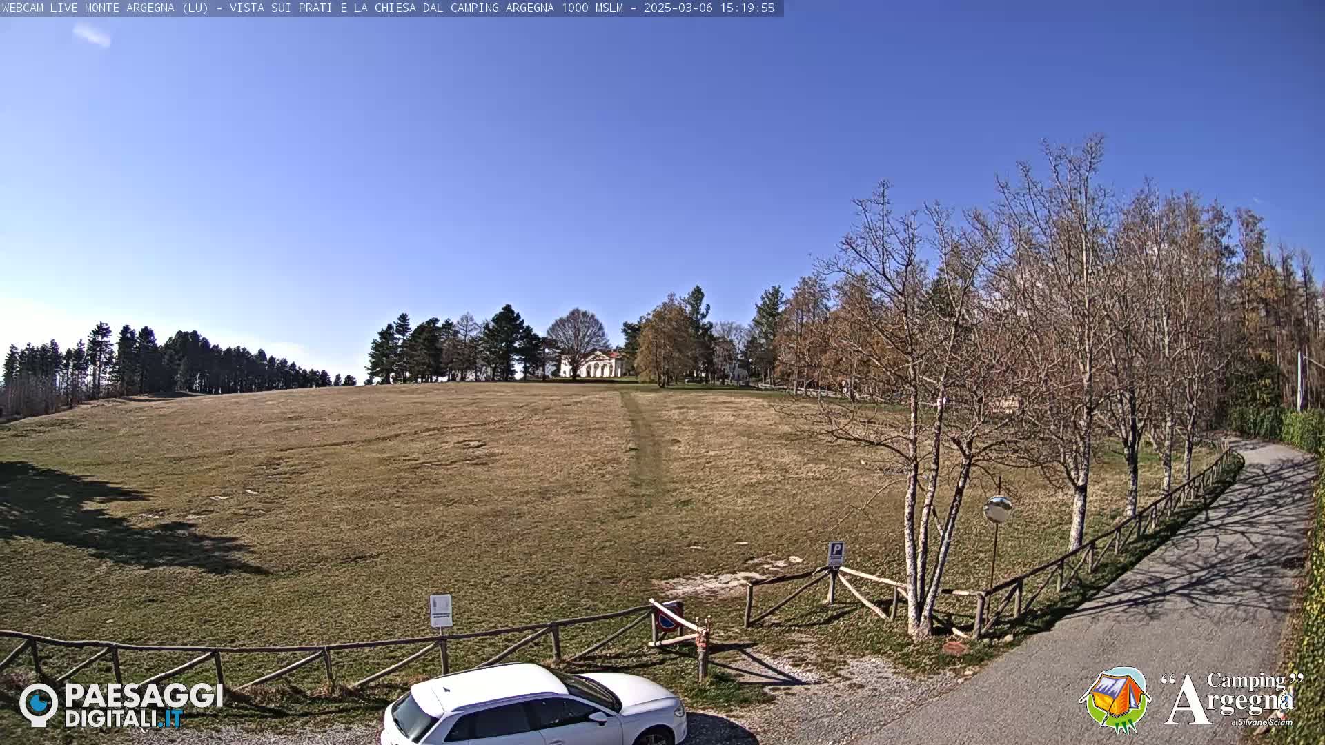 A white car is parked near a wooden fence bordering a grassy field with a building in the distance, under a clear blue sky.