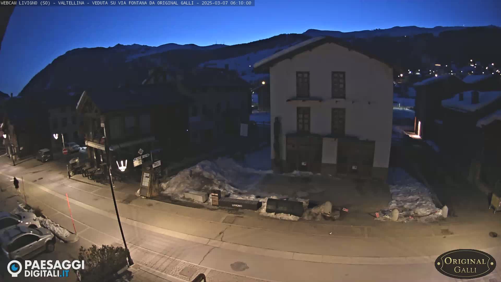 A nighttime street scene in a snow-covered village, showing several buildings under a dark blue, pre-dawn sky.