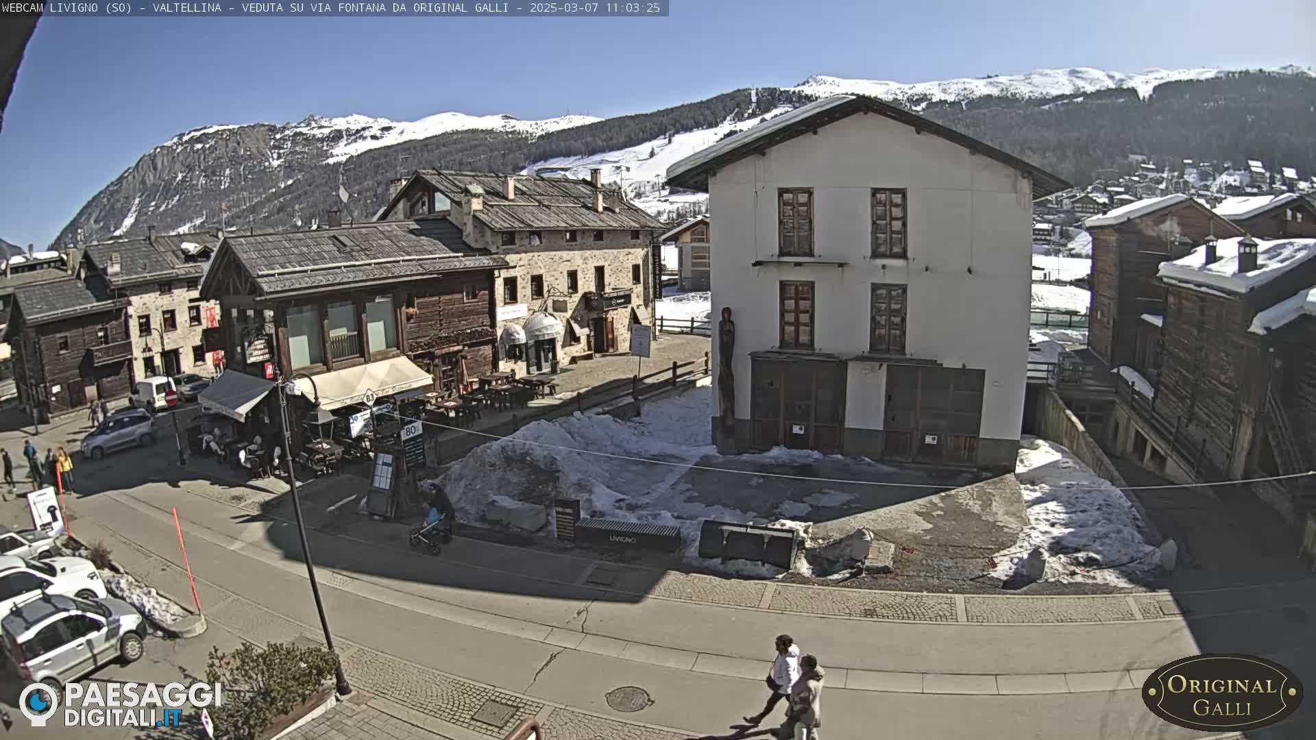 A sunny, snow-dusted village square in a mountainous region, with several buildings and a few pedestrians.