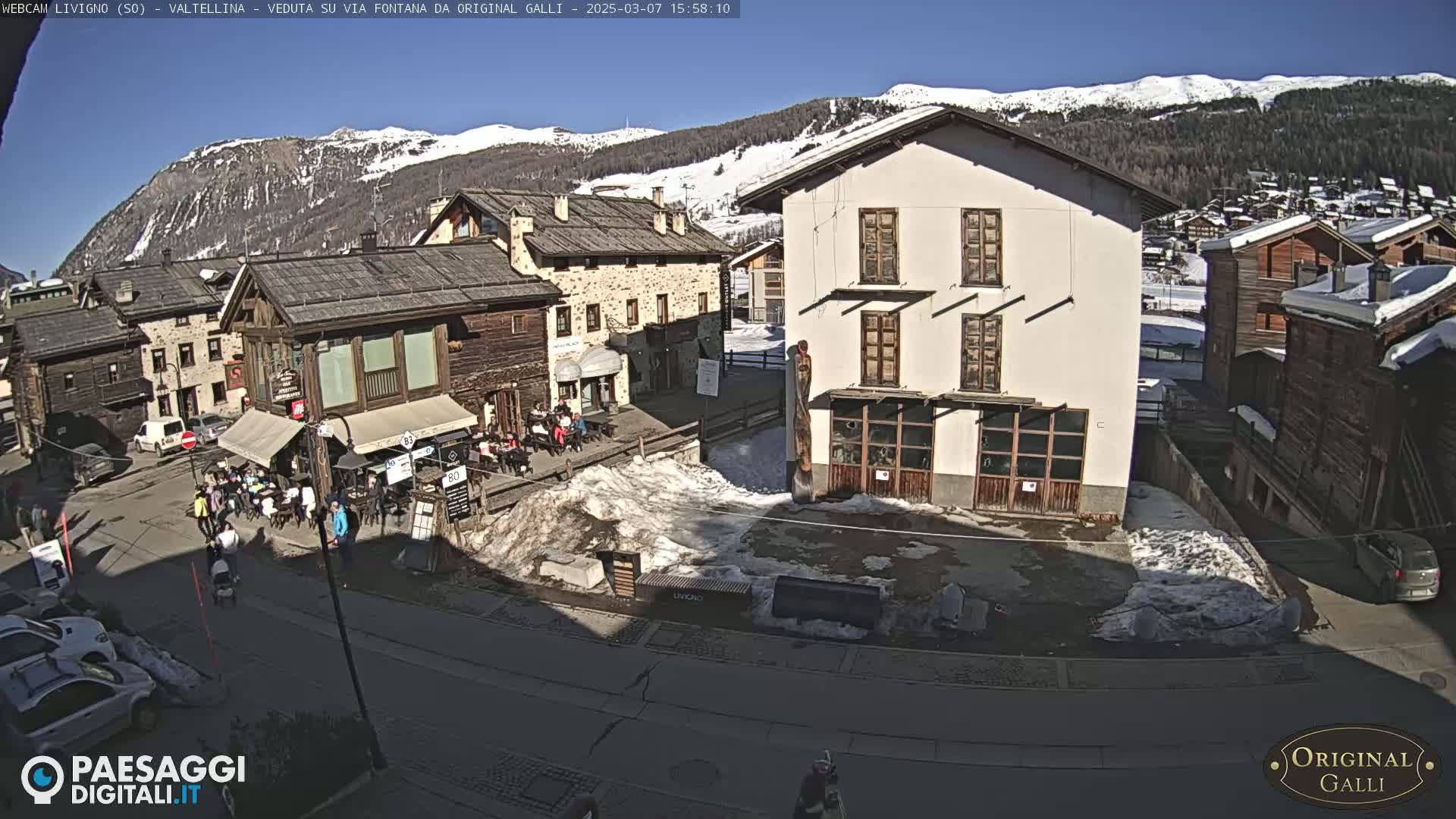 A sunny day in a snow-dusted village square with several buildings, some people, and cars parked nearby, mountains in the background.
