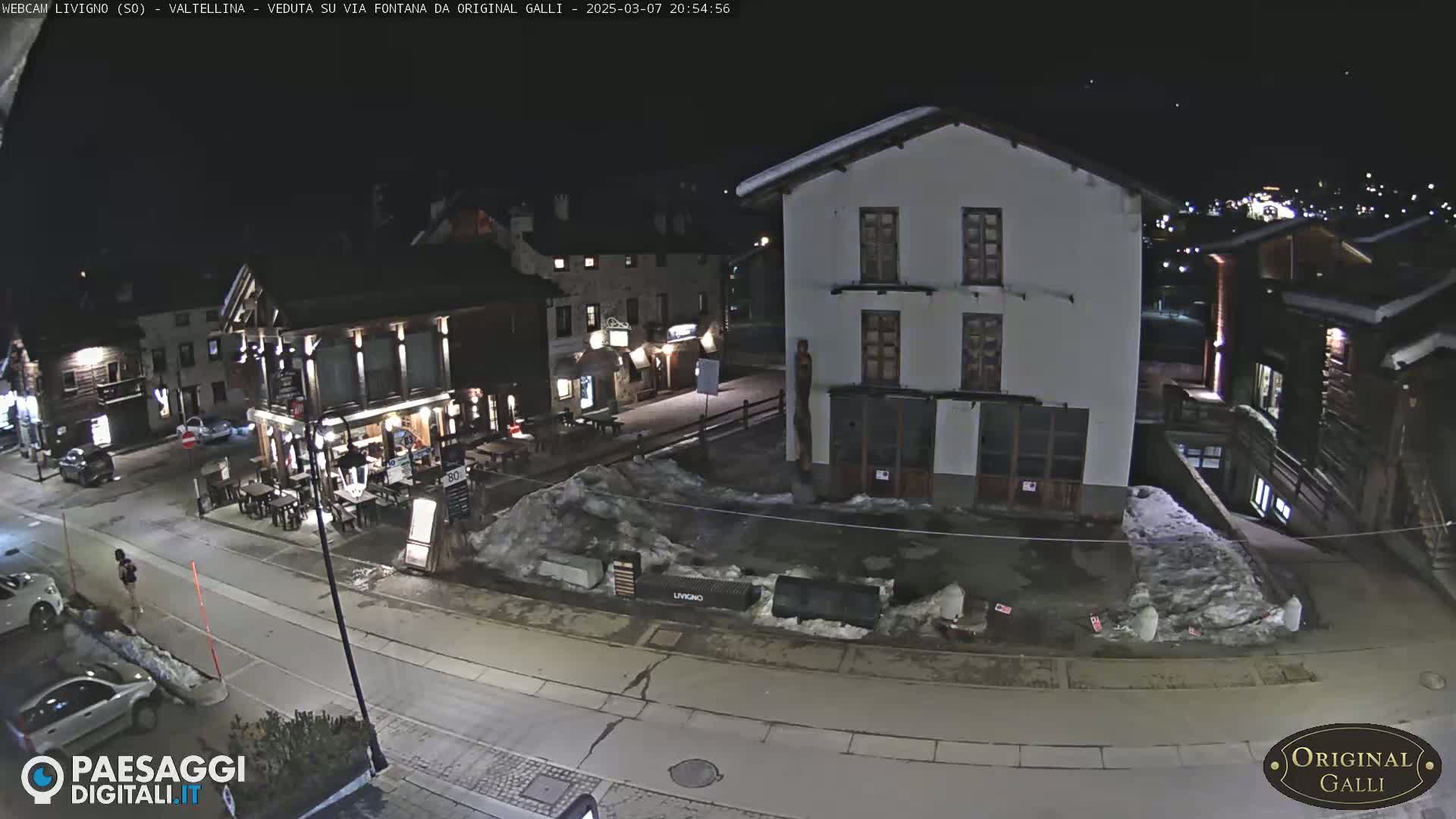 A nighttime, snow-covered town square with several buildings, cars, and a few people visible under dark skies.