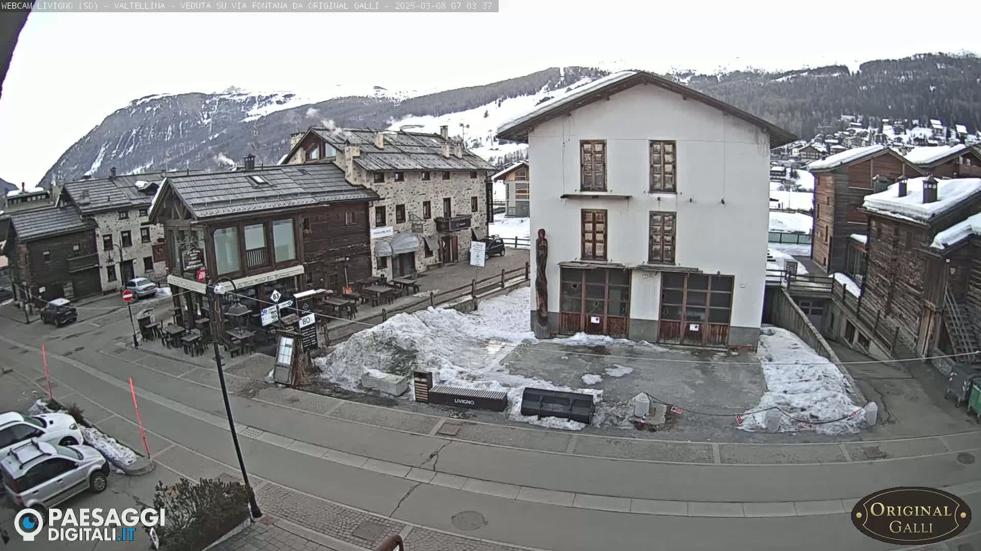 A snow-covered village square with several buildings, some cars parked, and patches of ice and snow on the ground, under an overcast sky.