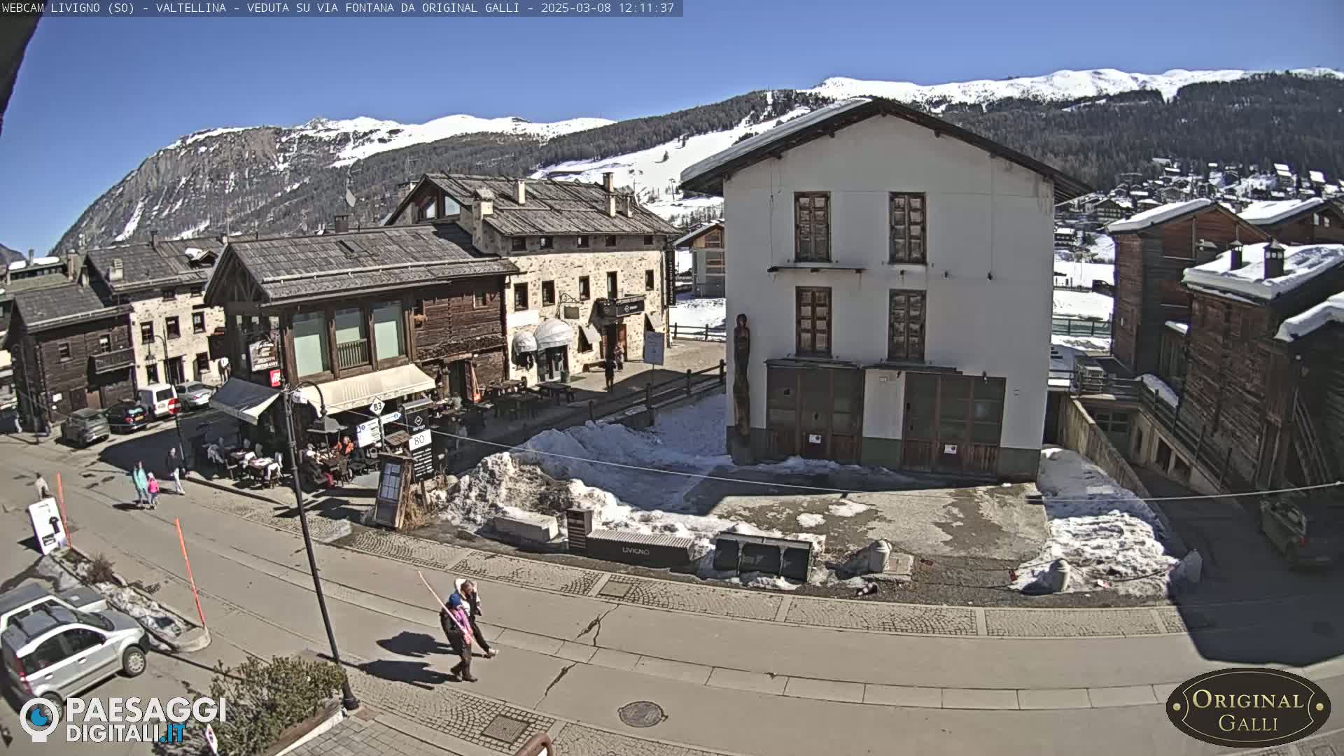 A sunny day in a snow-dusted village square, with several buildings, people walking, and a mountain range in the background.