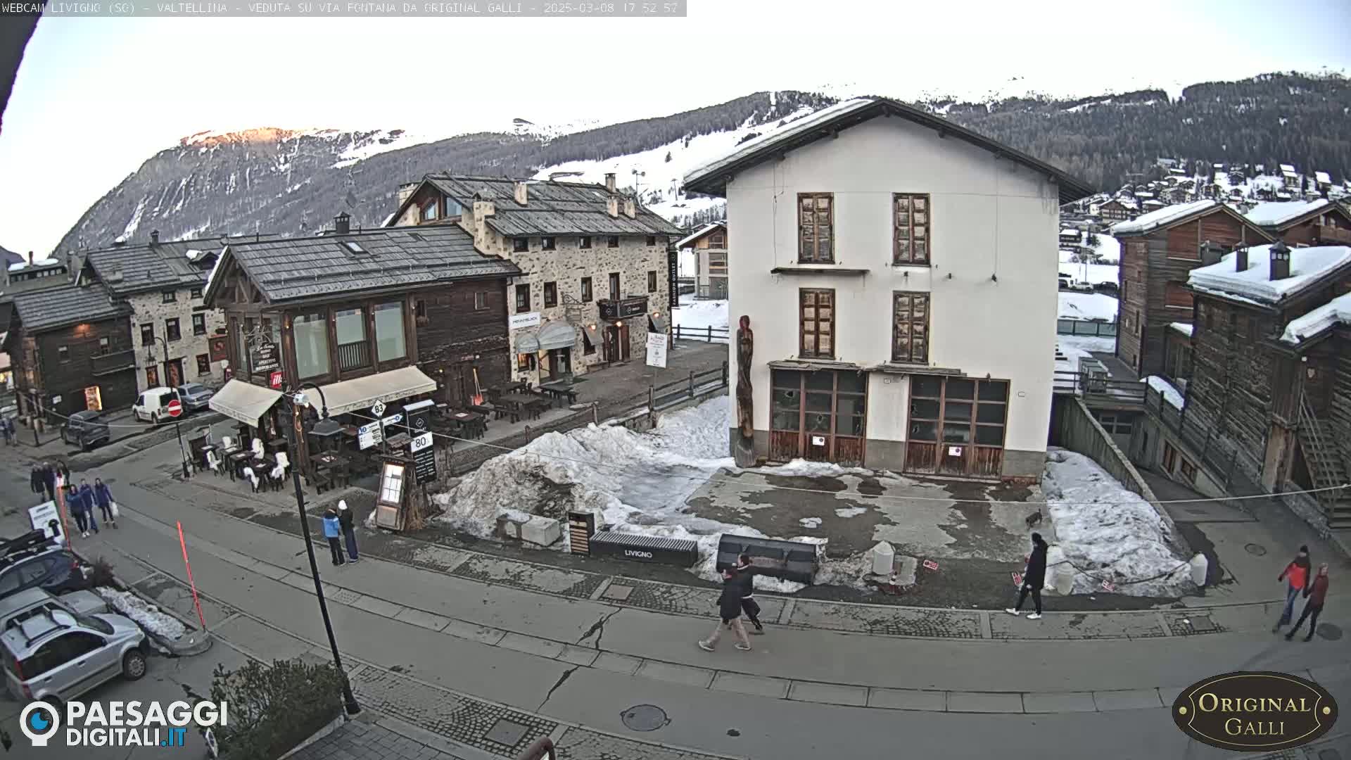 A snowy village square with several buildings, some people walking, and cars parked nearby, under a mostly clear sky.