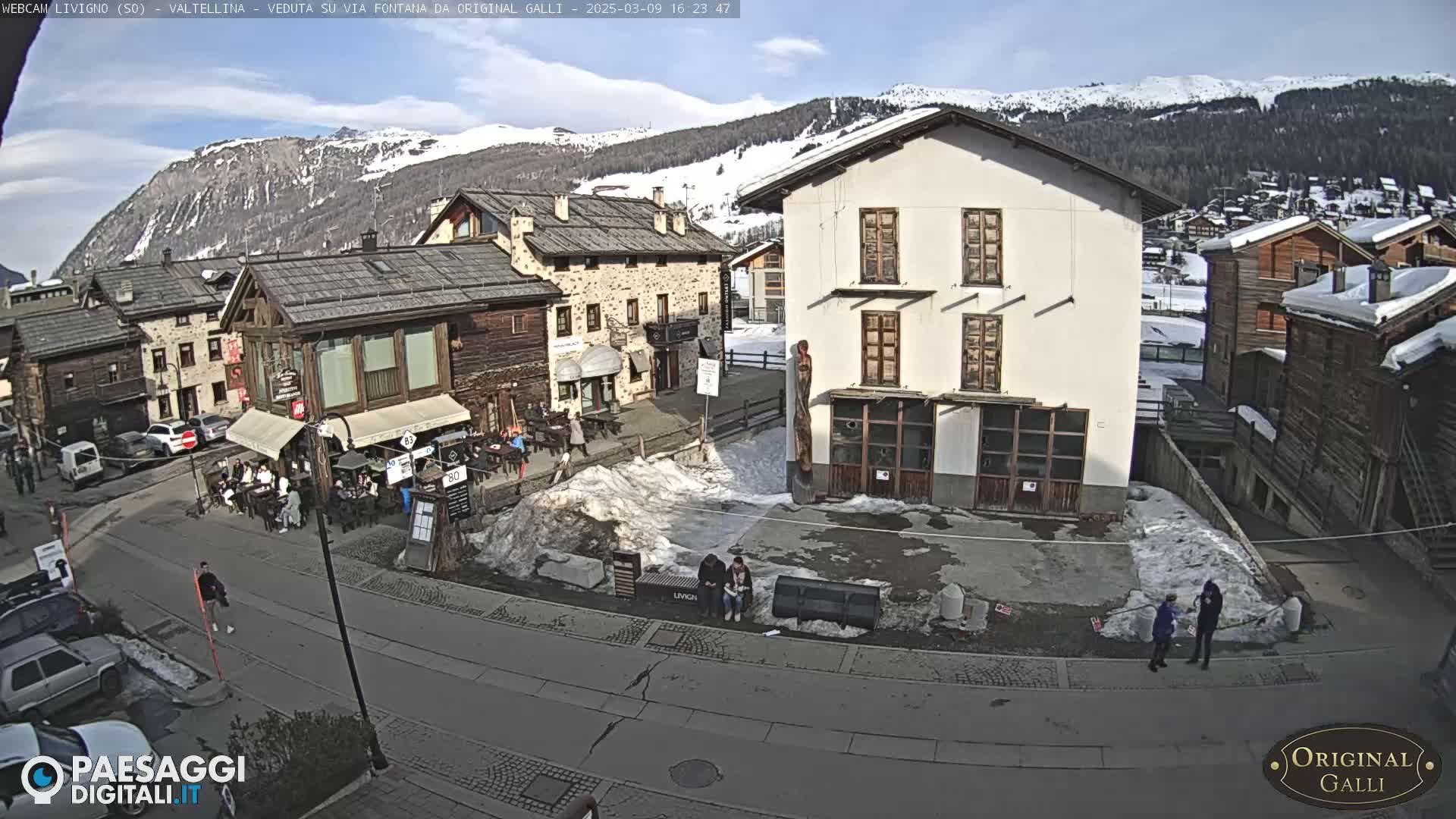 A sunny mountain village square with snow patches, several buildings, and a few pedestrians.