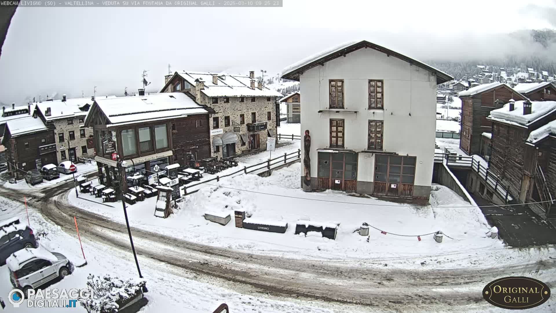 A snow-covered village square with several buildings, some cars parked, and outdoor seating areas.