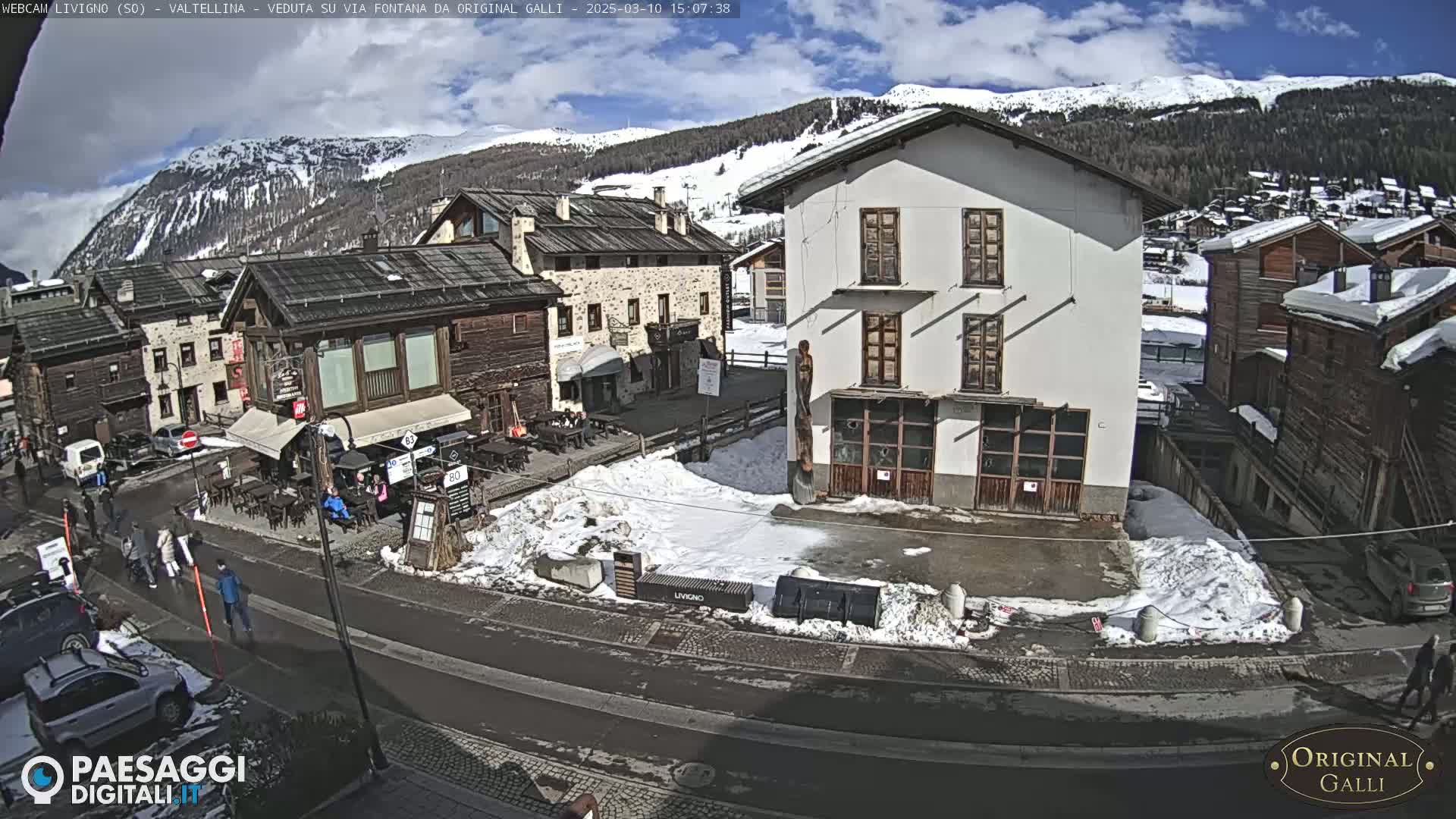 A partly sunny day in a snow-dusted village nestled in a mountain valley, showing a street scene with several buildings and a few pedestrians.