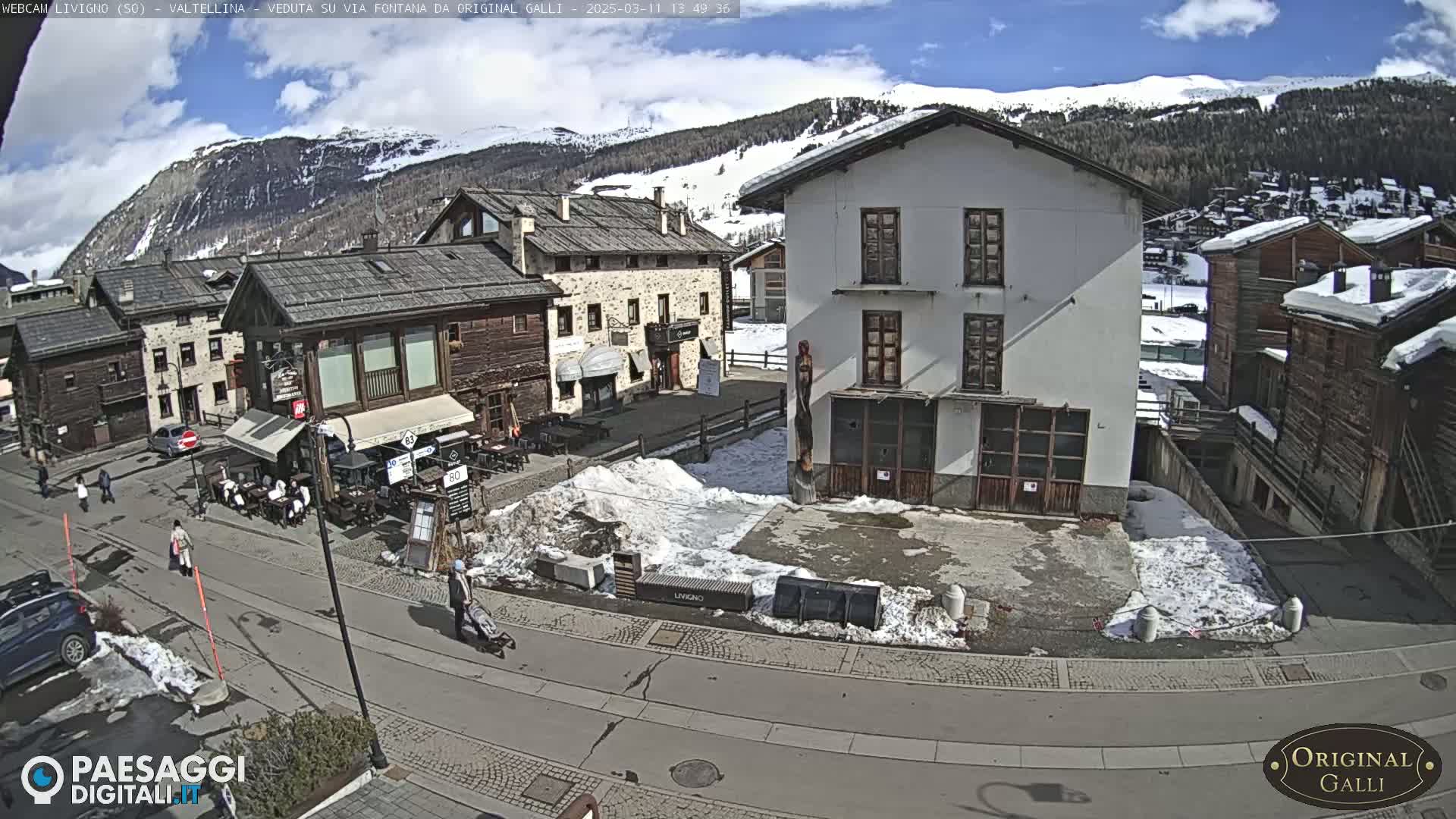 A sunny day in a snow-dusted mountain village square with several buildings, some people walking, and cars parked.