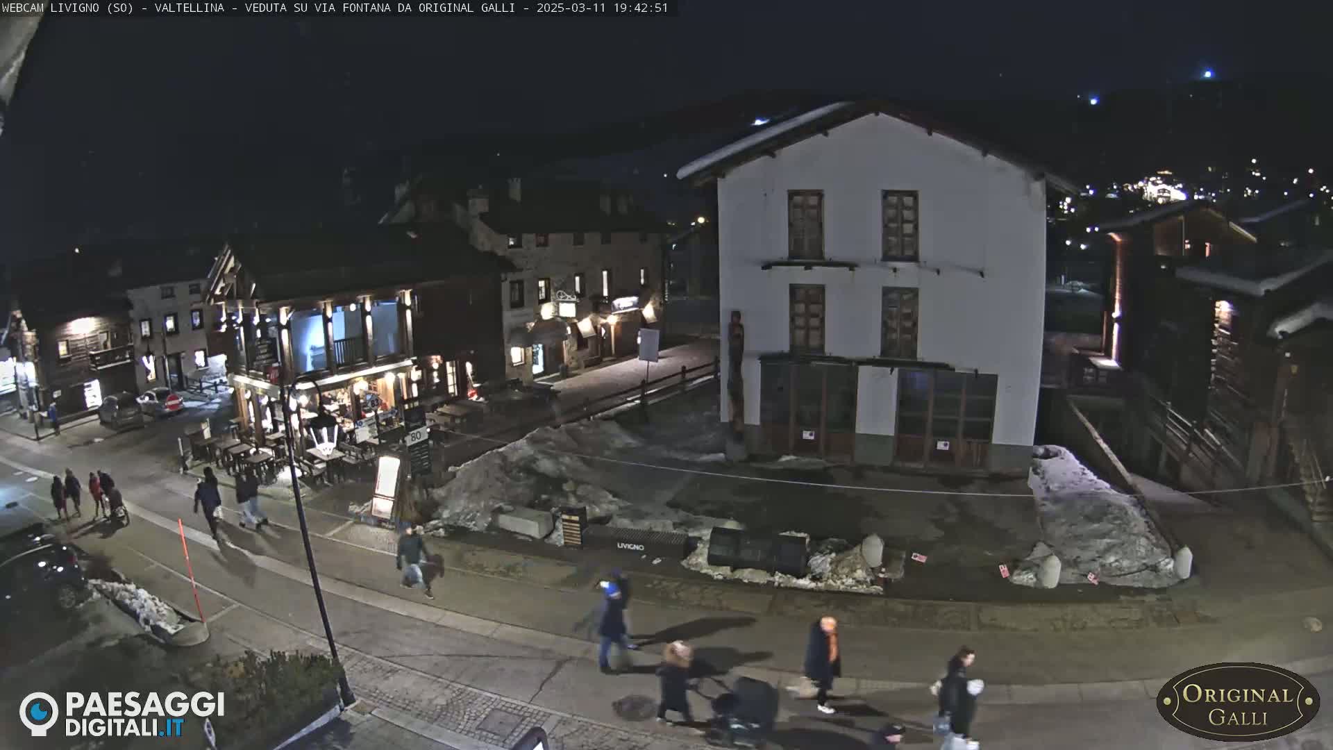 A nighttime street scene in a snow-dusted village shows several pedestrians walking past buildings and a restaurant, under a dark sky.