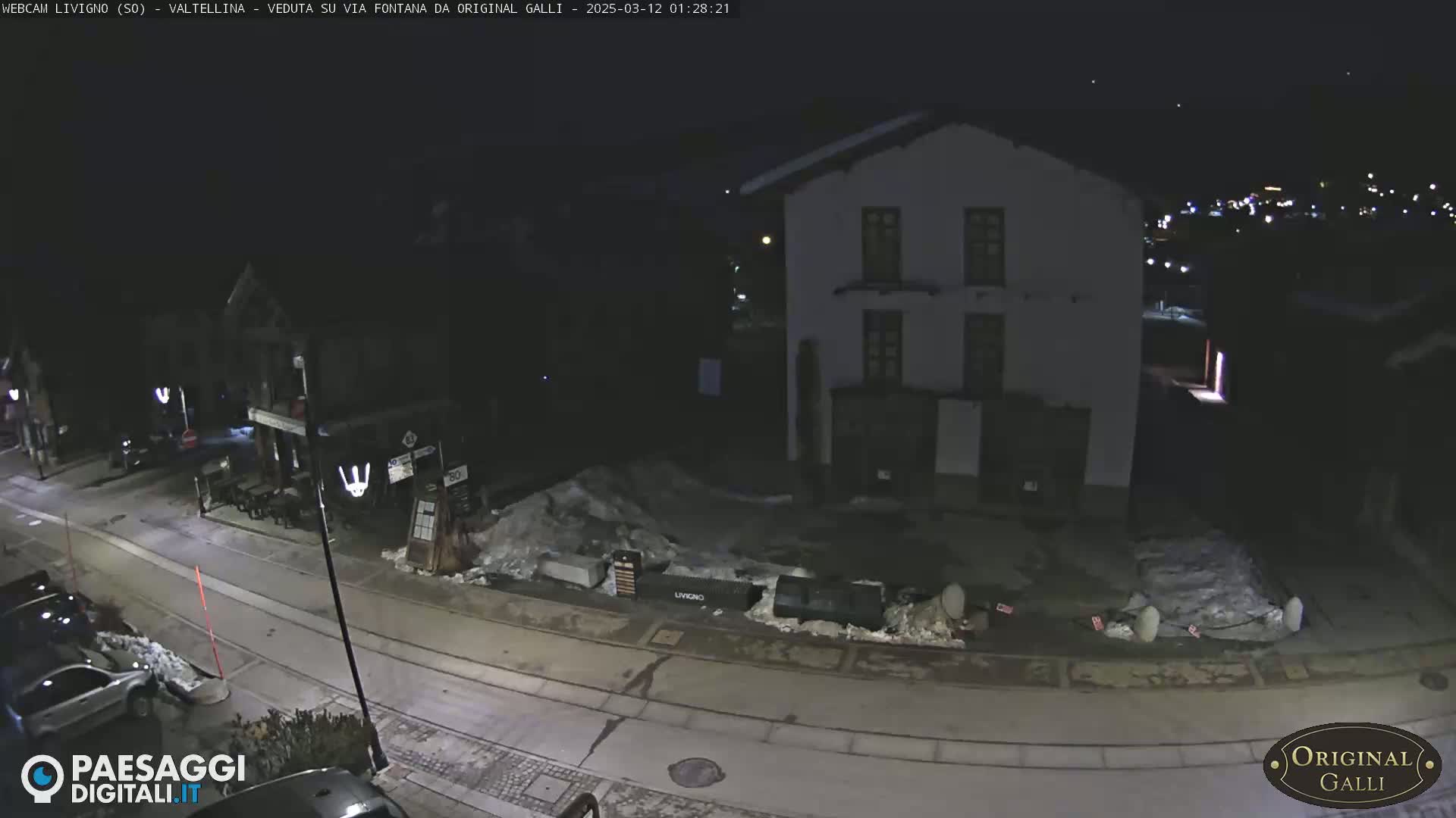 A nighttime street scene shows a snow-covered town square with a few parked cars and a two-story building under a dark, clear sky.