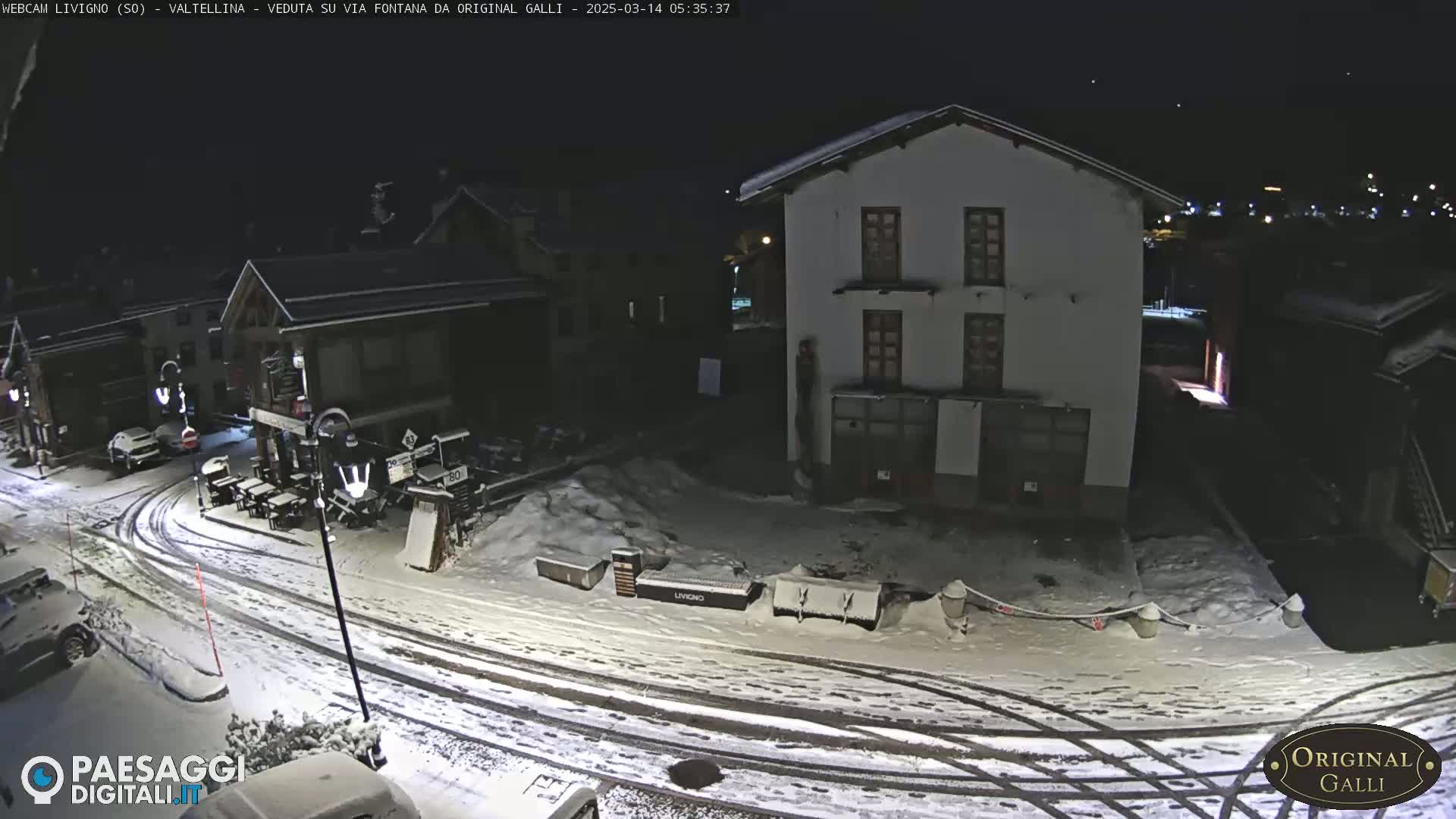 A snow-covered street scene at night shows several buildings, parked cars, and tire tracks in the snow.