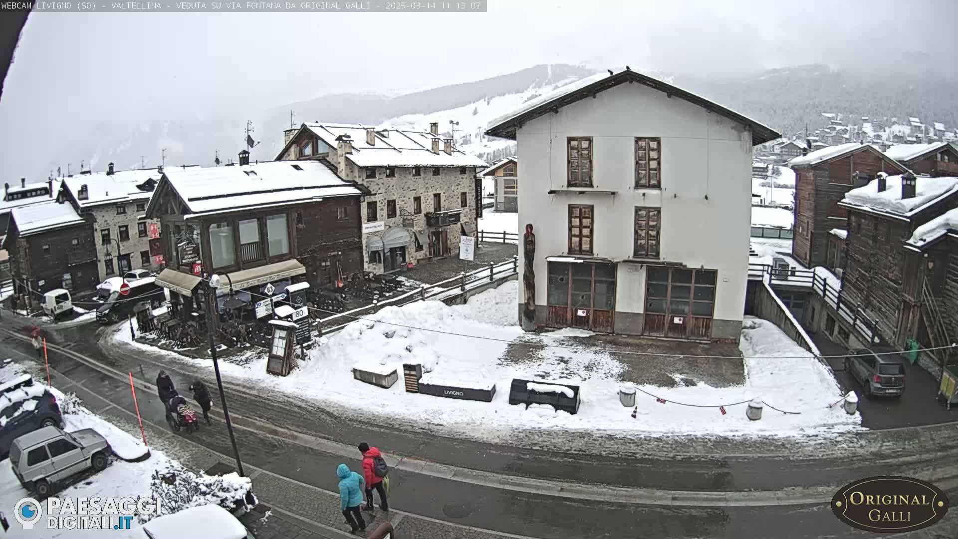 A snow-covered village square with several buildings, a few people walking, and cars parked along a street in overcast weather.