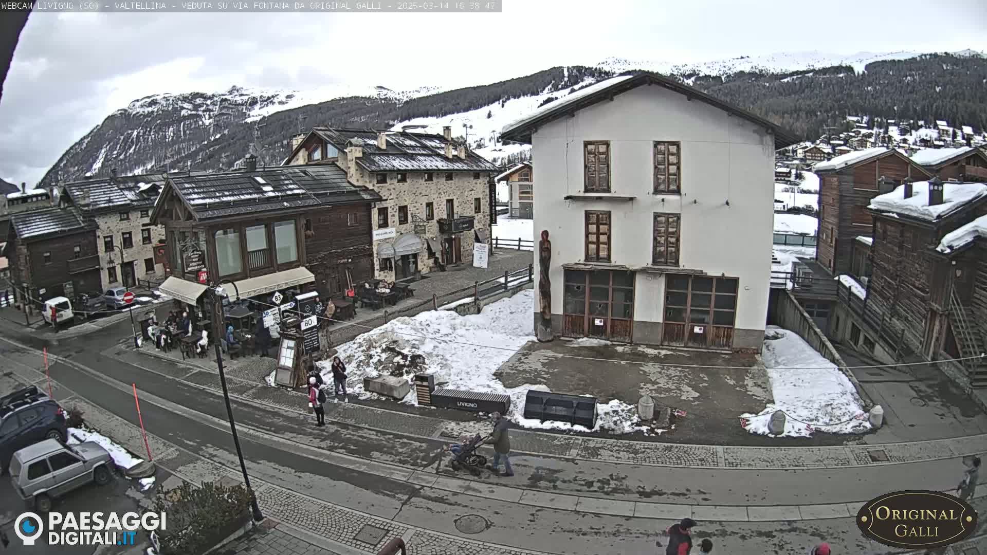 A snow-dusted village square with several buildings, a few pedestrians, and cars parked along a street, all under a cloudy sky.