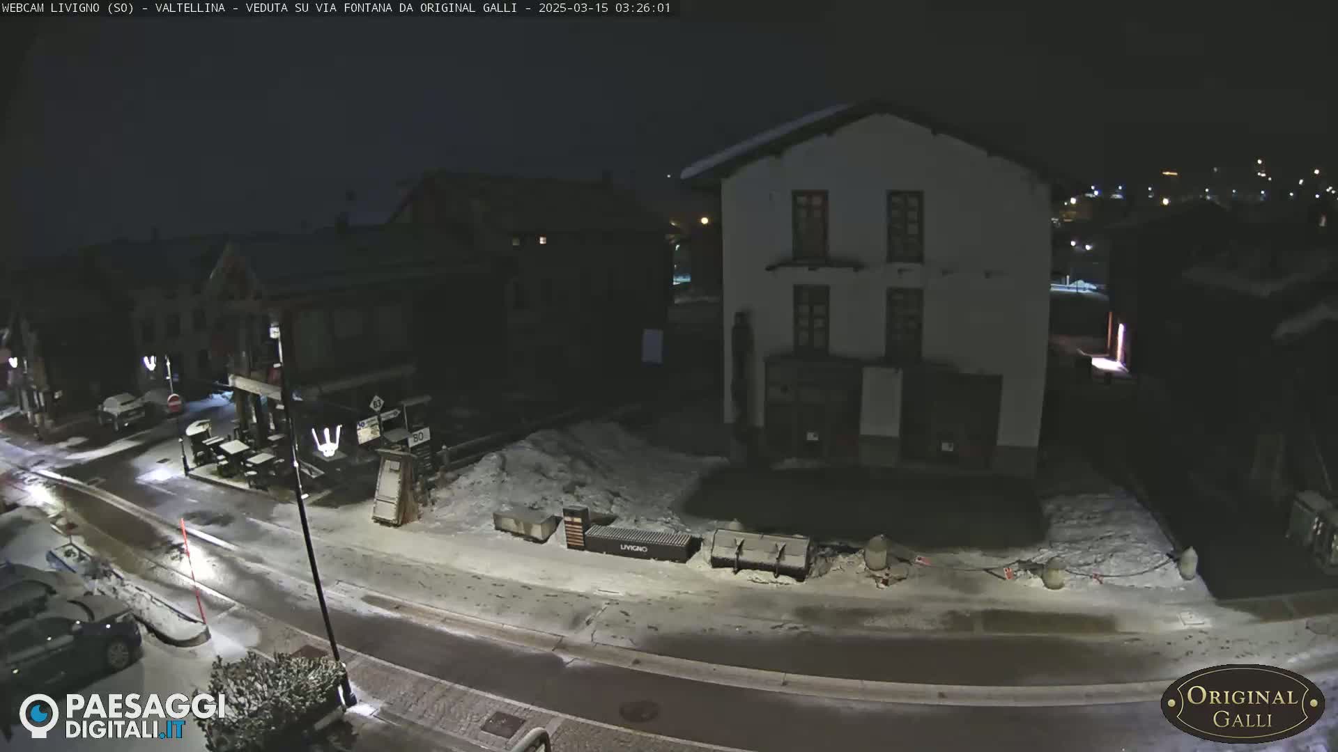 A snow-covered street at night shows several buildings and parked cars under dim lighting.