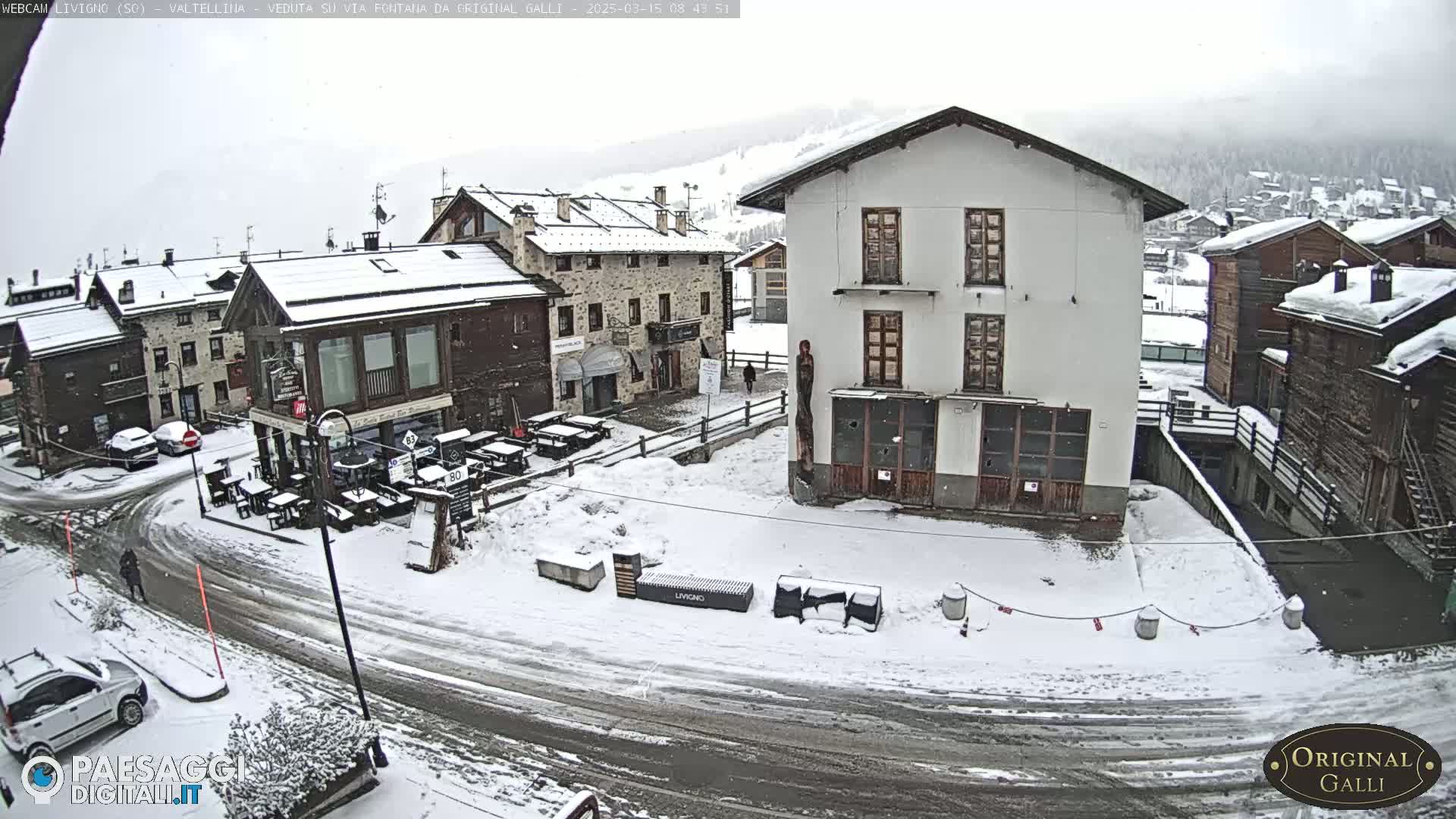 A snow-covered village street scene shows several buildings, some with snow-covered roofs and outdoor seating areas, under a lightly snowing sky.