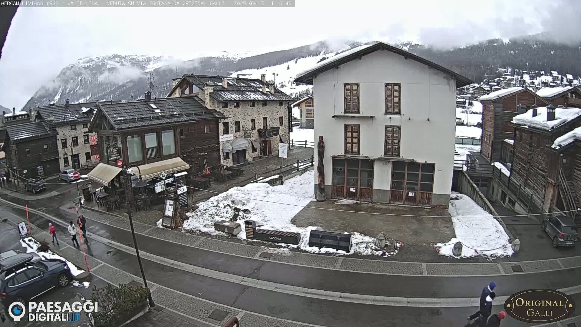 A snow-covered village street scene shows several pedestrians walking past stone and wood buildings under an overcast sky.
