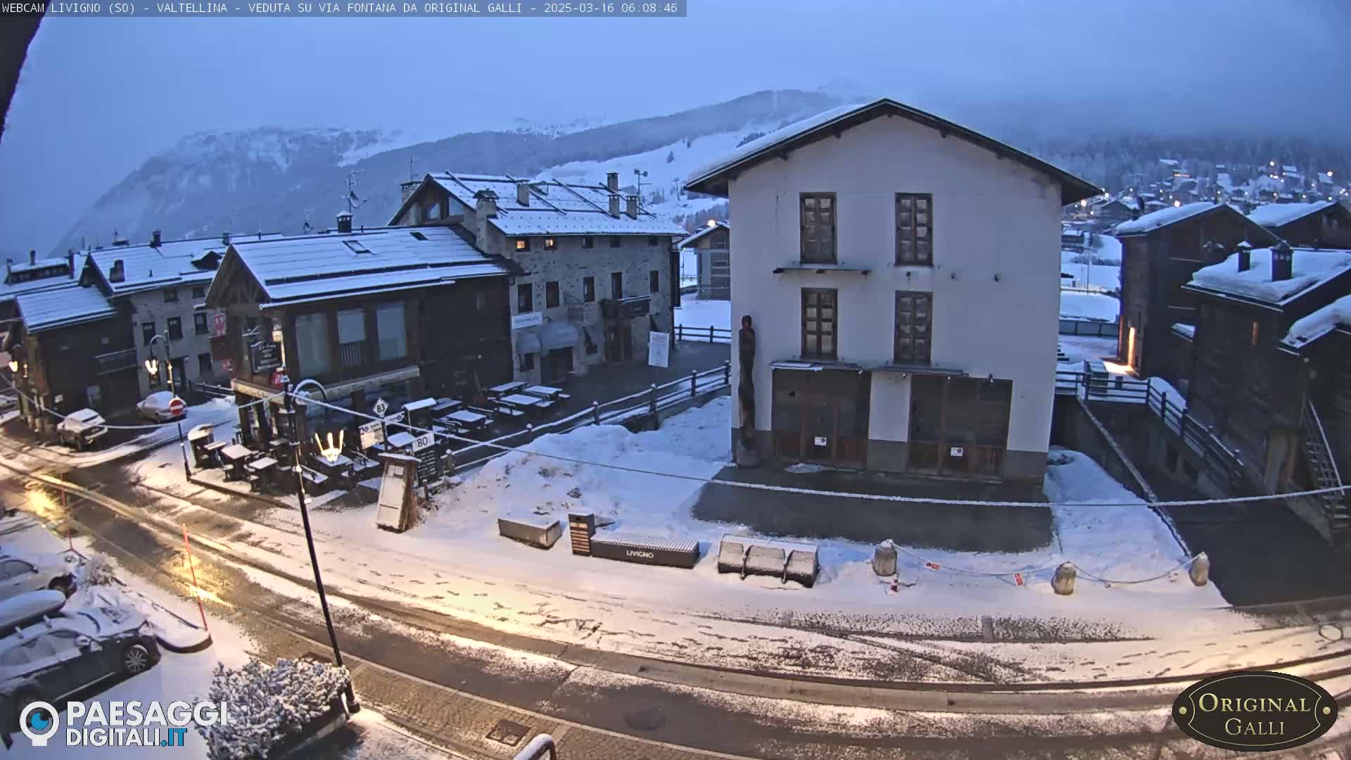 A snow-covered village street scene shows several buildings, parked cars, and outdoor seating areas under a gray, overcast sky.