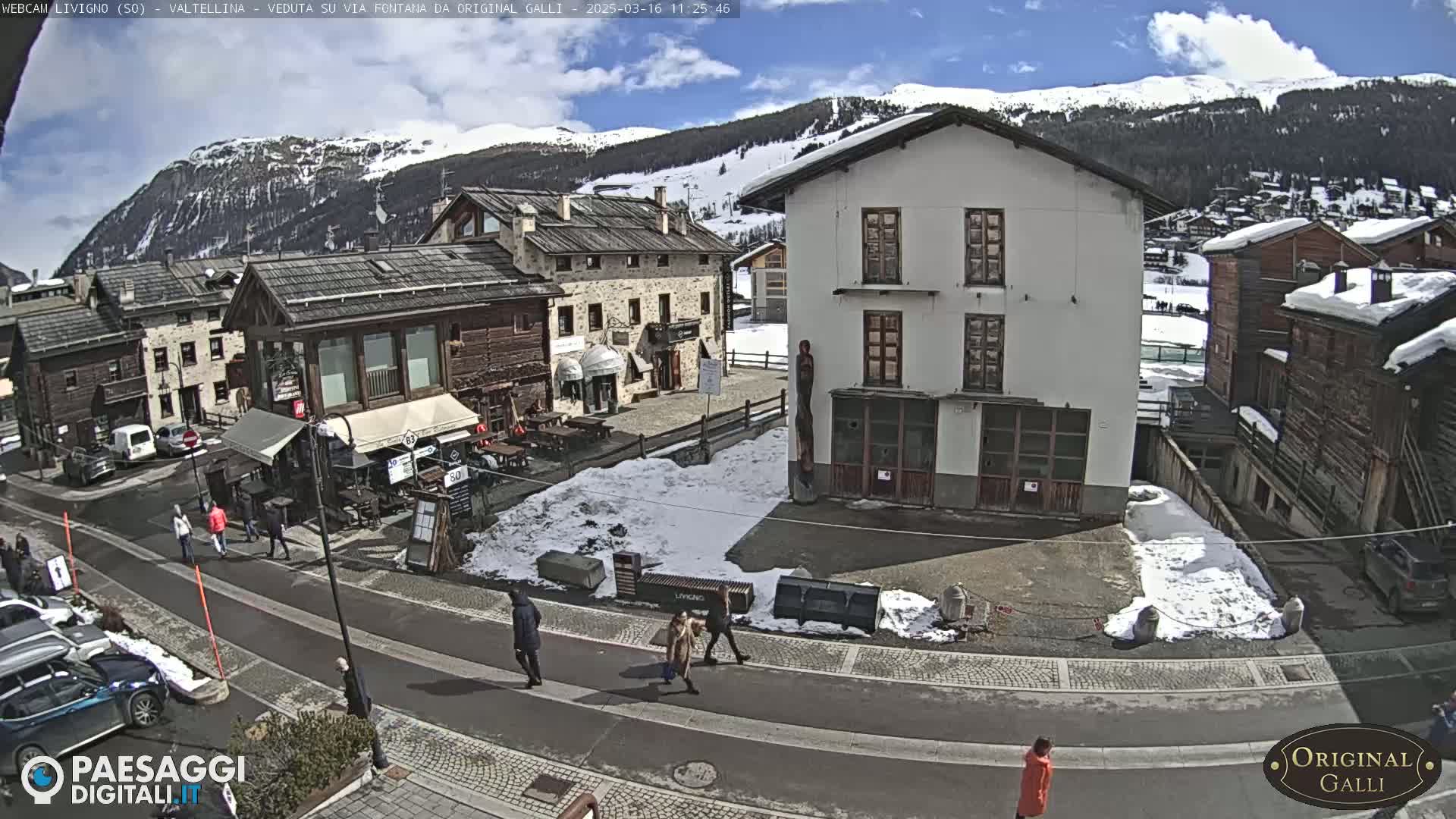 A street scene in a snow-dusted mountain village on a partly sunny day shows pedestrians walking past buildings and parked cars.