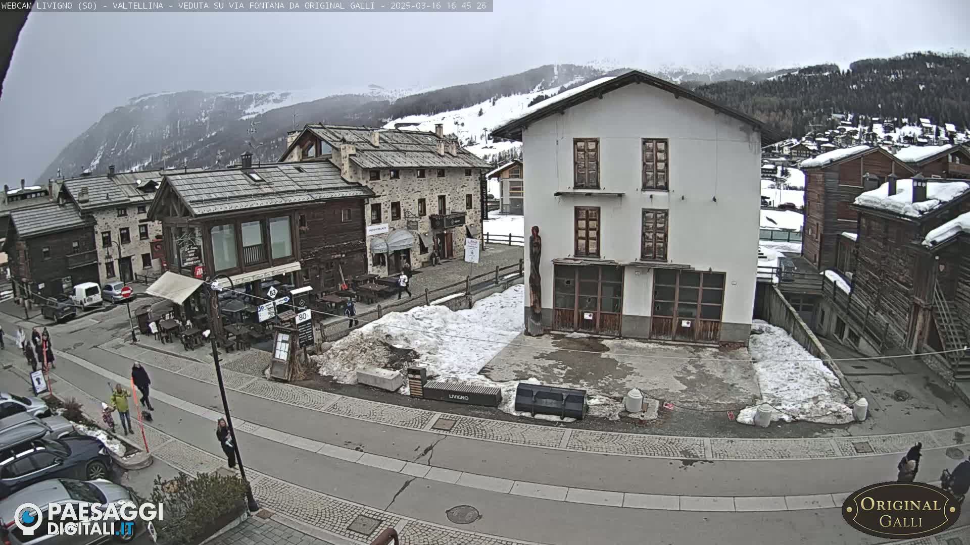 A village street scene with several buildings, some snow on the ground, and a few pedestrians, under an overcast sky.