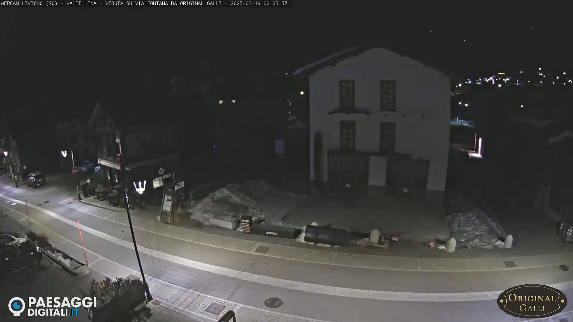 A nighttime street scene shows a mostly empty road, a few parked cars, a building, and piles of snow or ice.