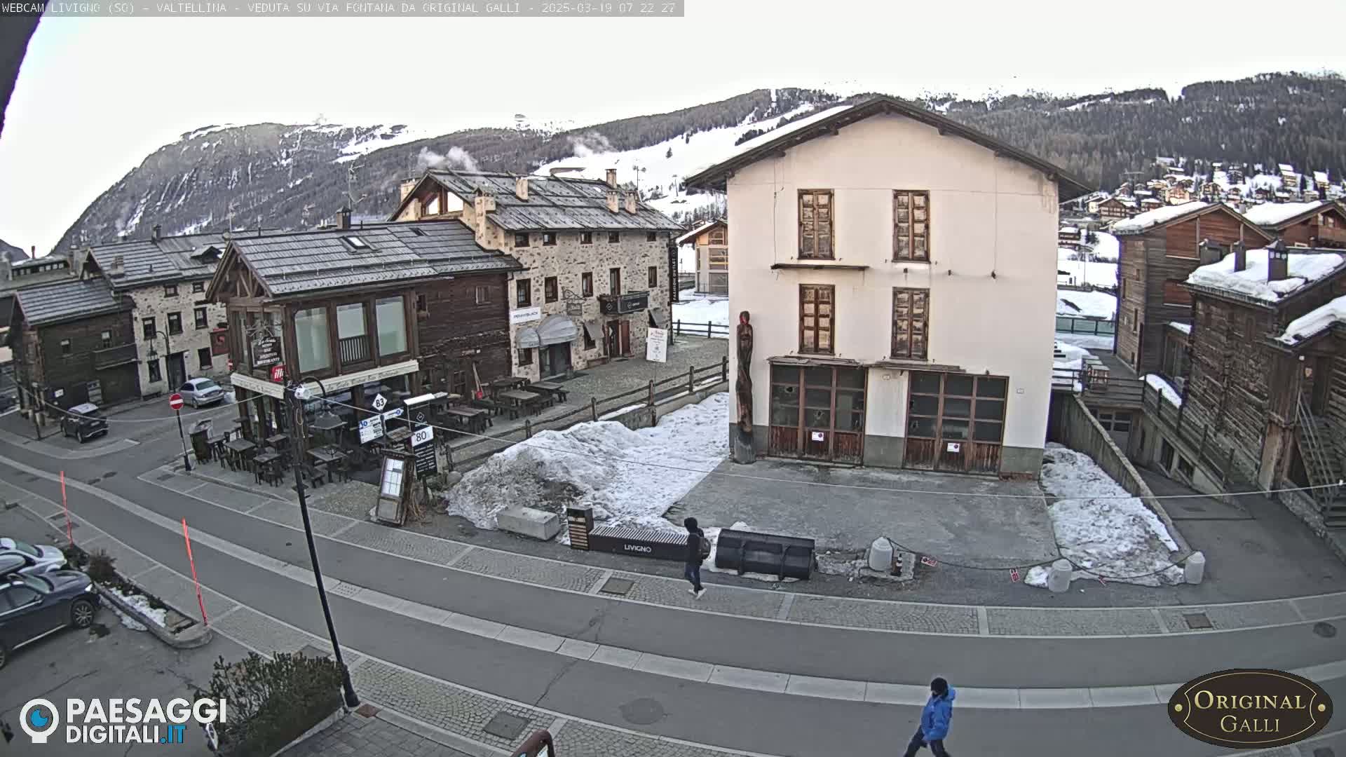A snow-dusted village square with several buildings, a few cars, and pedestrians, all under a mostly cloudy sky.