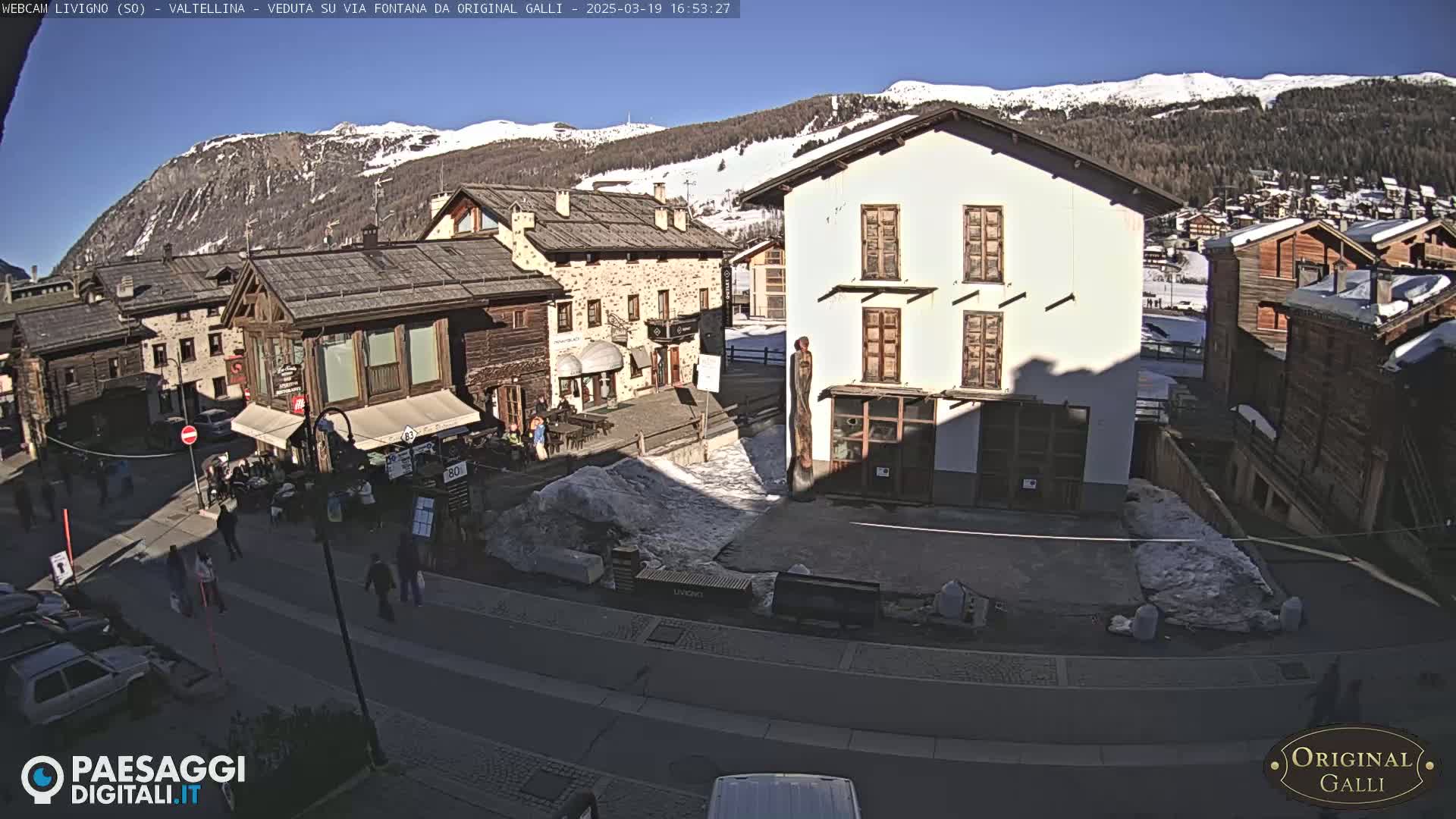 A sunny day in a snow-dusted mountain village shows a street scene with several buildings, some people walking, and cars parked along the road.