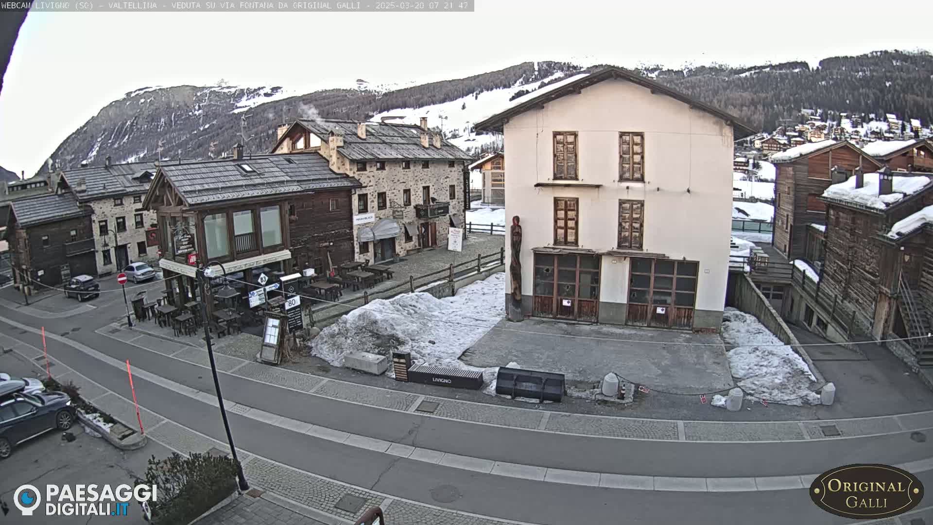 A snow-dusted village square in a mountainous region, with several buildings and cars visible under a partly cloudy sky.
