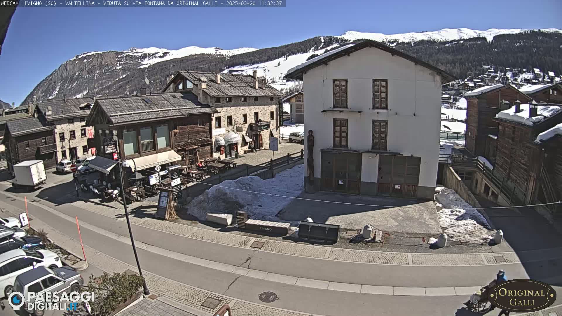 A sunny day in a snow-dusted village square surrounded by stone and wood buildings, with mountains in the background.