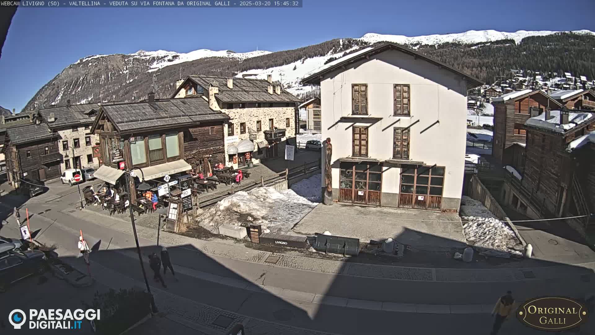 A sunny, snow-dusted village square is viewed from above, showing several buildings, some with outdoor seating, and a few pedestrians.