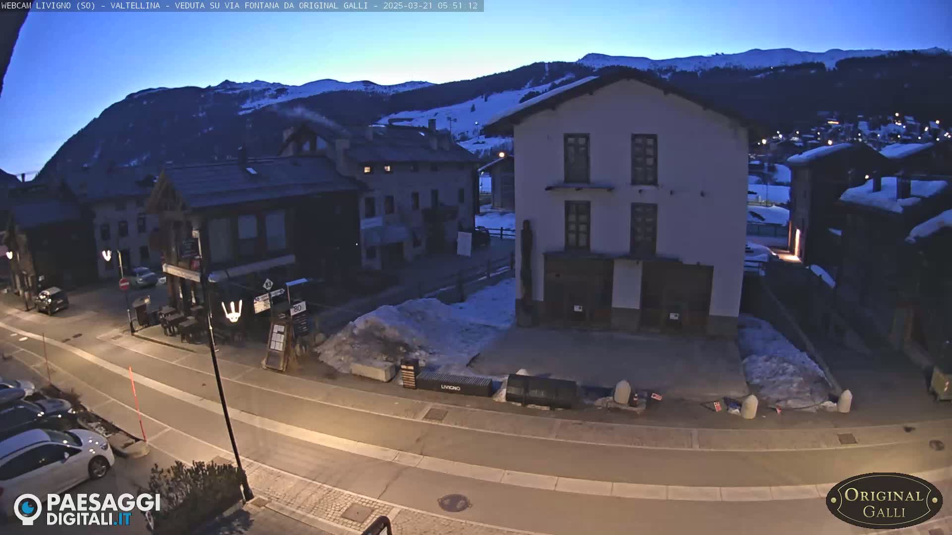 A twilight view of a snow-dusted village street with several buildings, some cars parked along the side of the road, and a mountain range in the background under a clear, dark blue sky.
