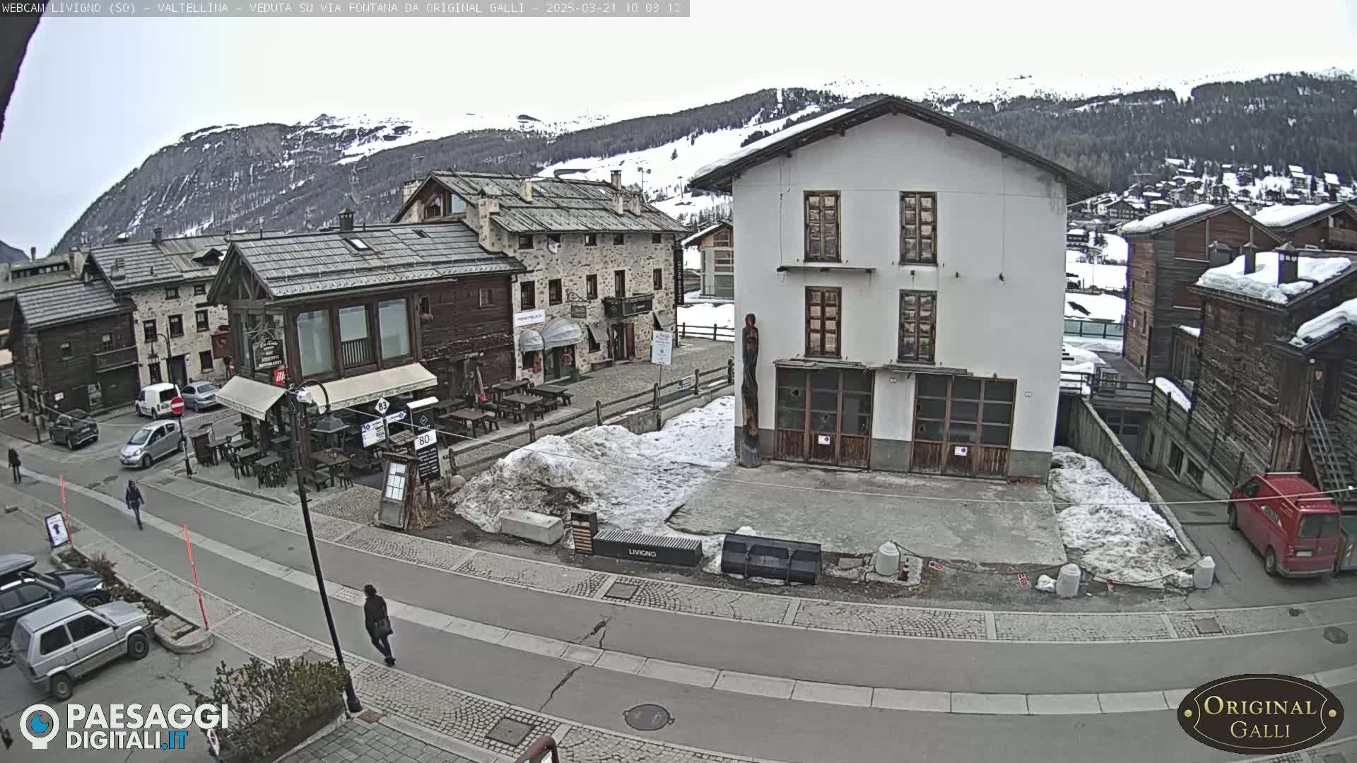 A snowy village square in a mountainous region, with several stone buildings and cars parked along a street, under an overcast sky.