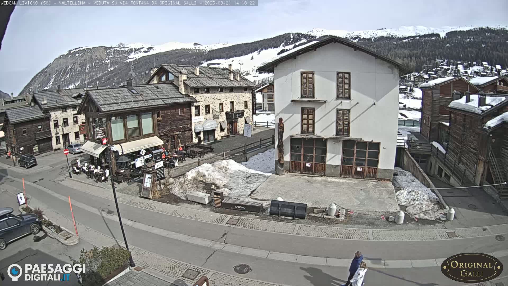 A sunny day in a snow-dusted village square shows several buildings, some with outdoor seating, a road, and a few pedestrians.