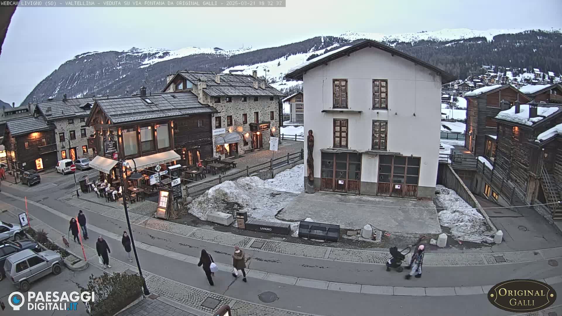 A snow-dusted village square in the mountains, with pedestrians walking on a street lined with buildings and a few parked cars under a twilight sky.