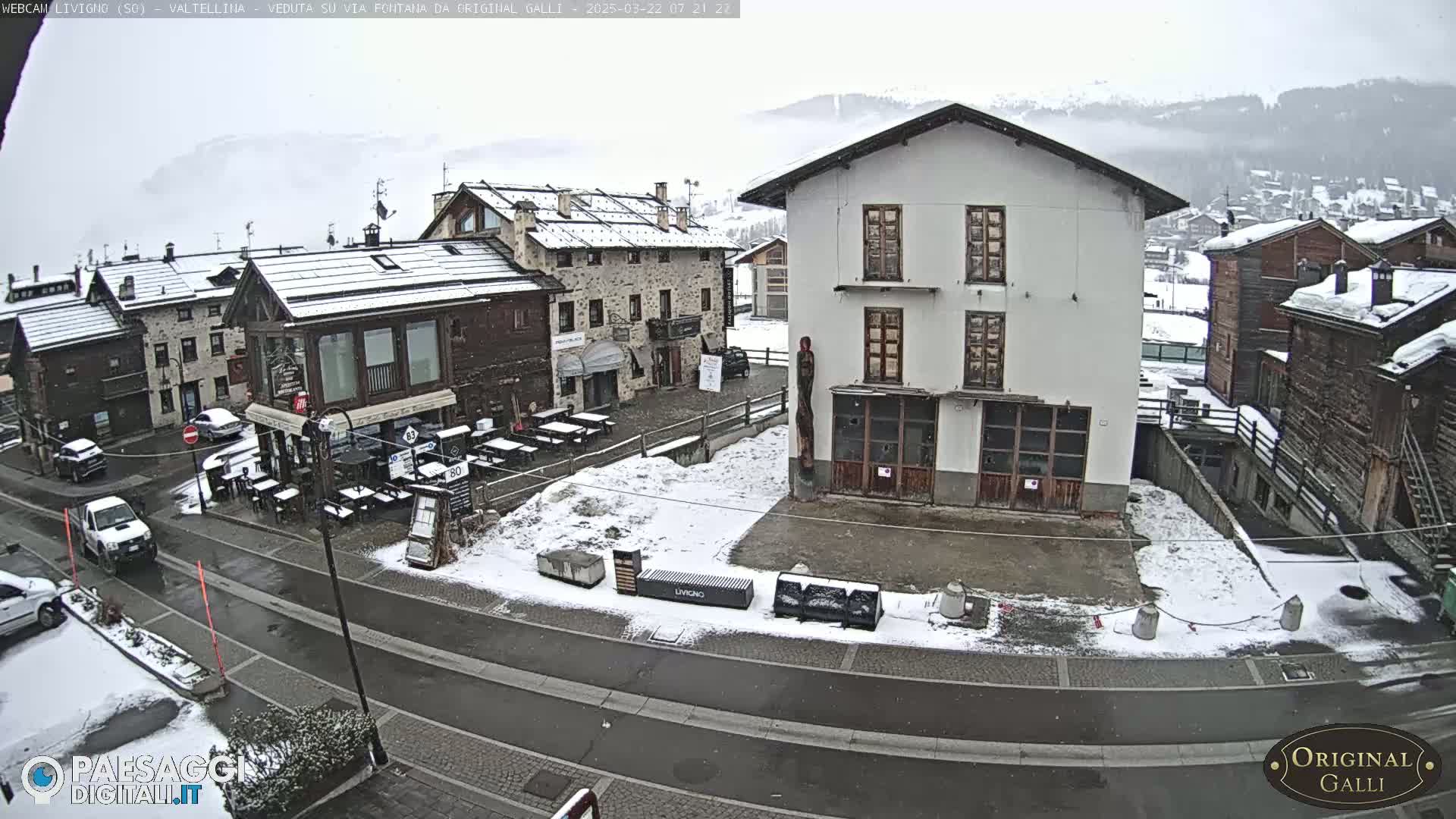 A snow-covered town square with several buildings and parked cars, under a lightly snowing sky.
