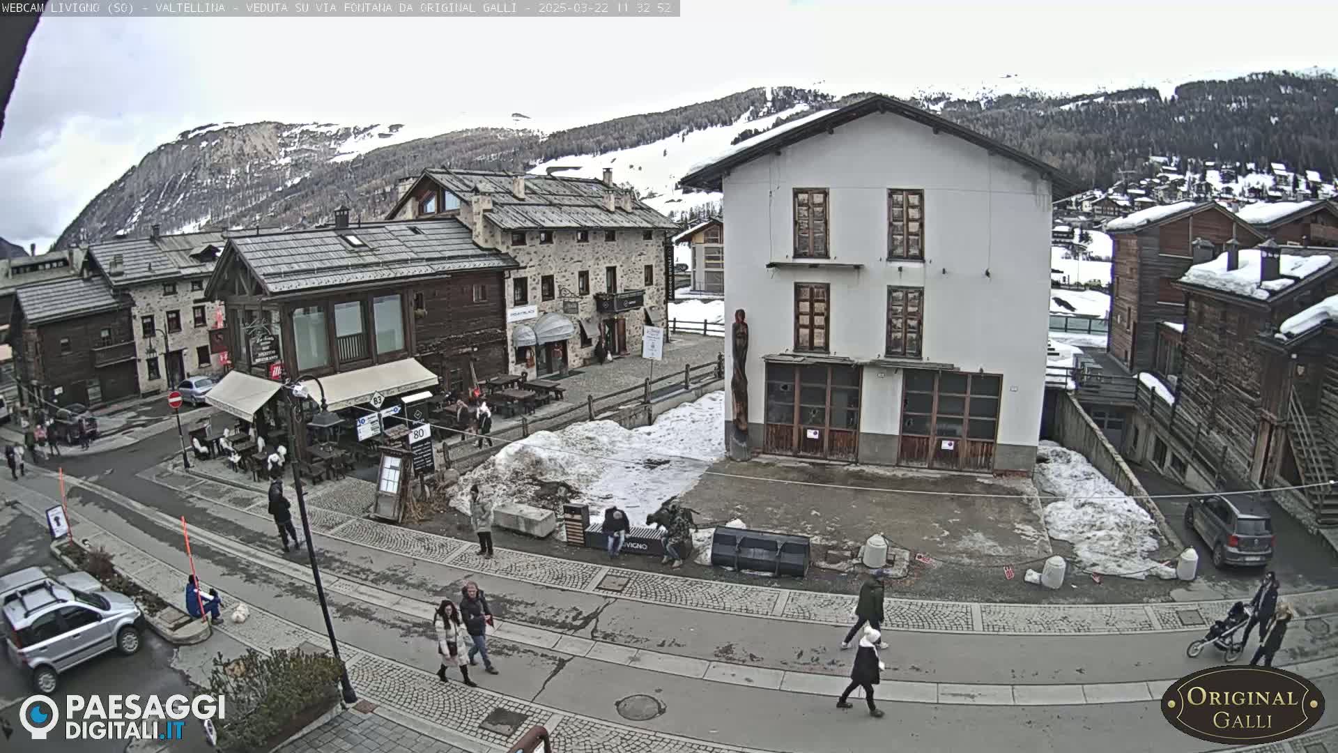 A mostly clear day in a snow-dusted village square, with pedestrians walking on streets flanked by stone buildings and a mountain range in the background.