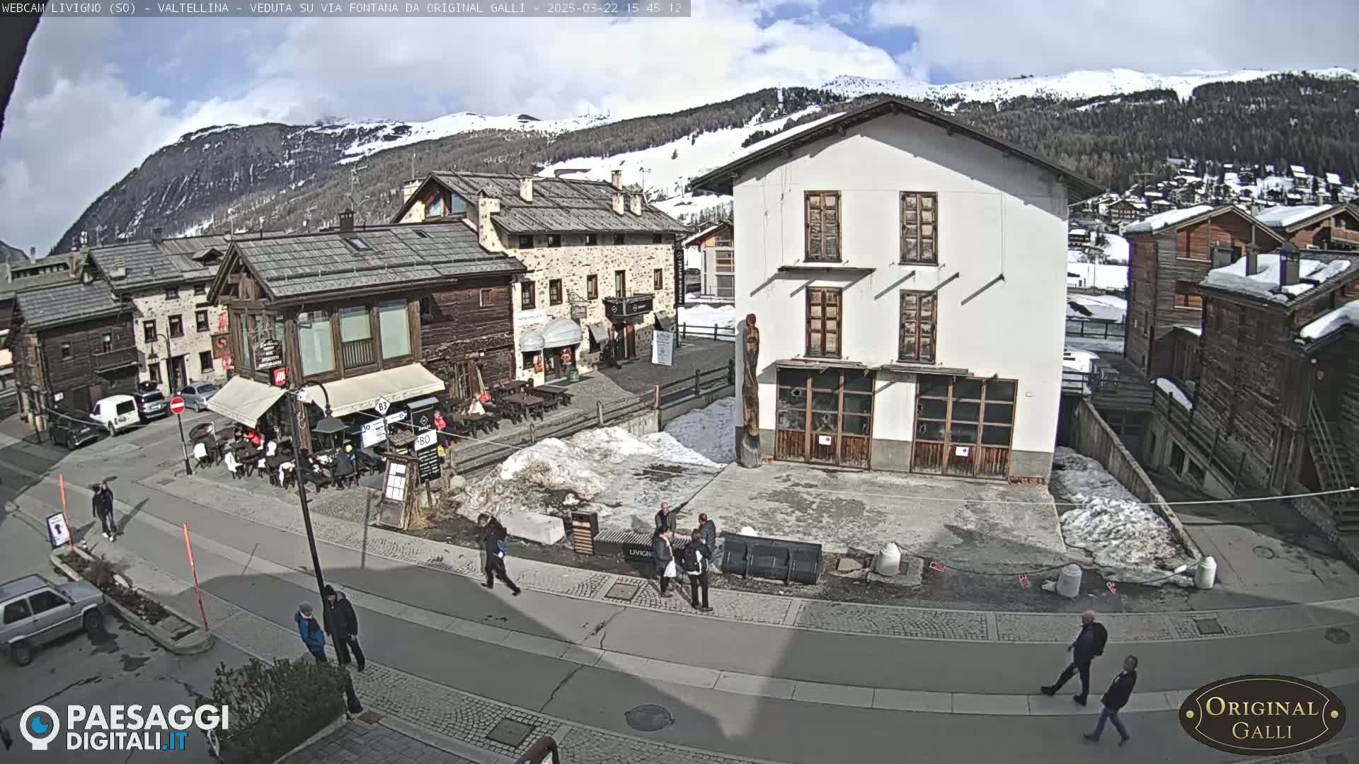 A sunny day in a snow-dusted mountain village square with several people walking among stone and wood buildings.