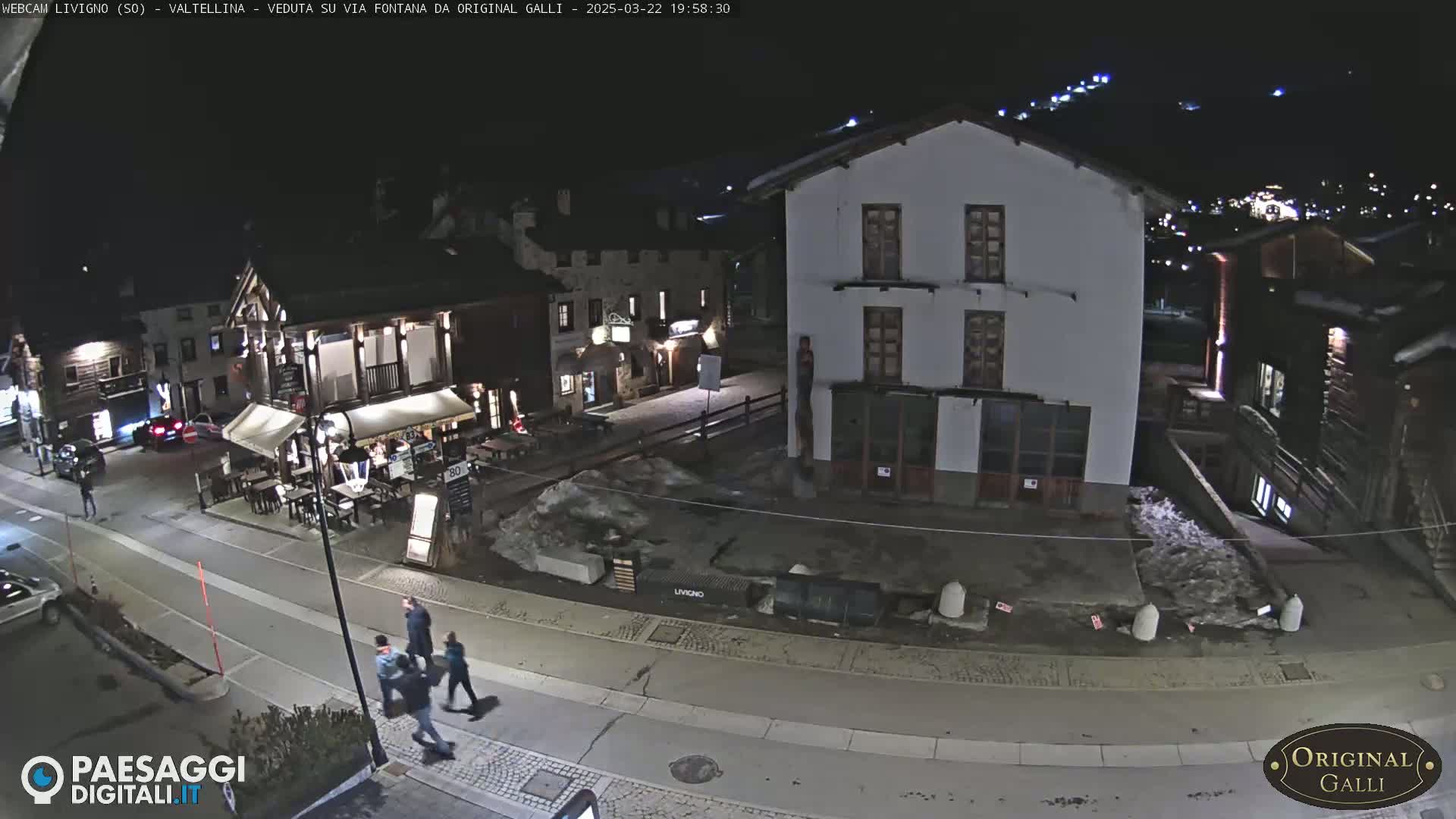 A nighttime street scene in a village, showing several buildings, a few people walking, and patches of snow or ice on the ground.