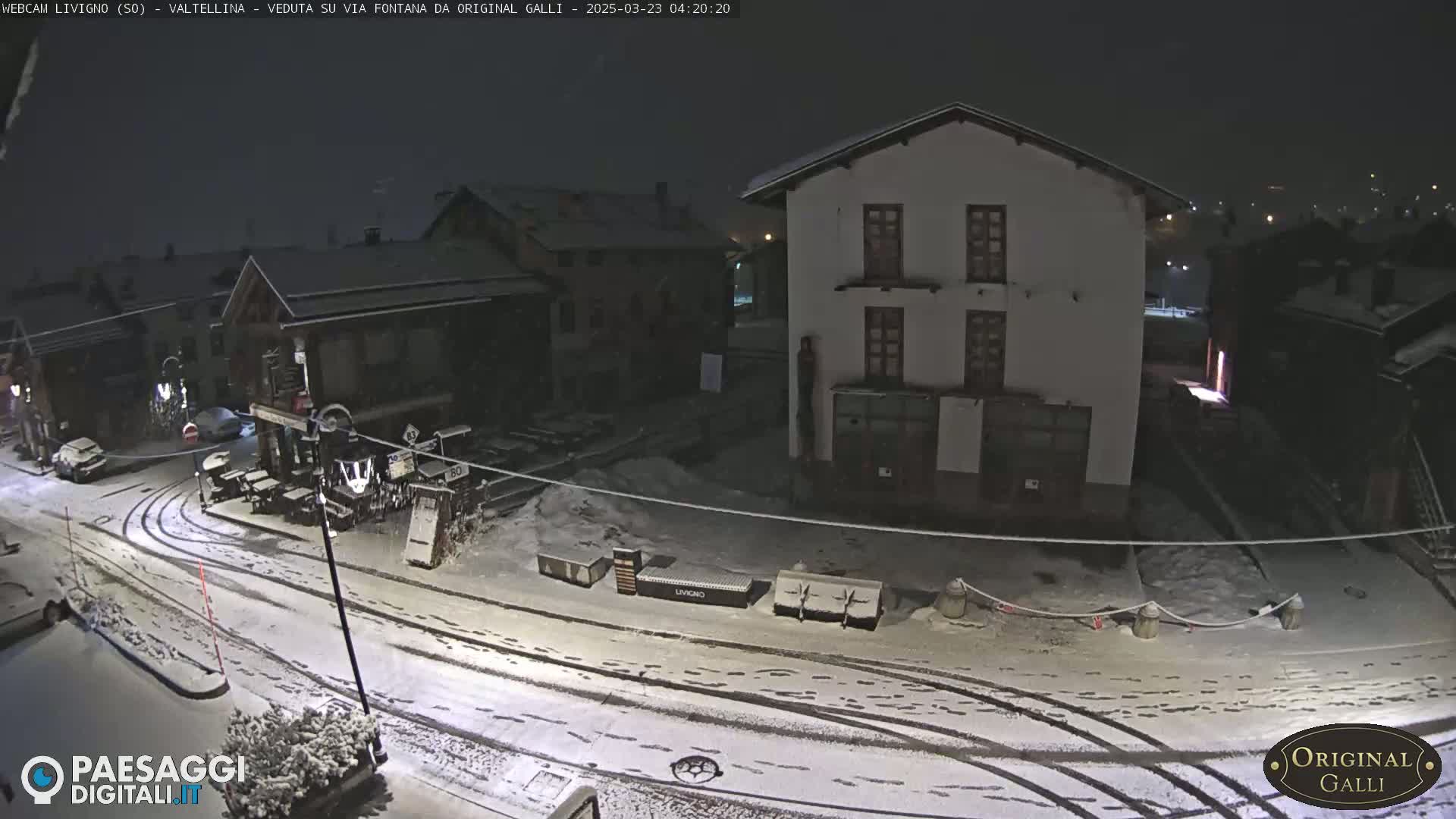 A snow-covered town square at night, with several buildings and a few parked cars visible under dim lighting.