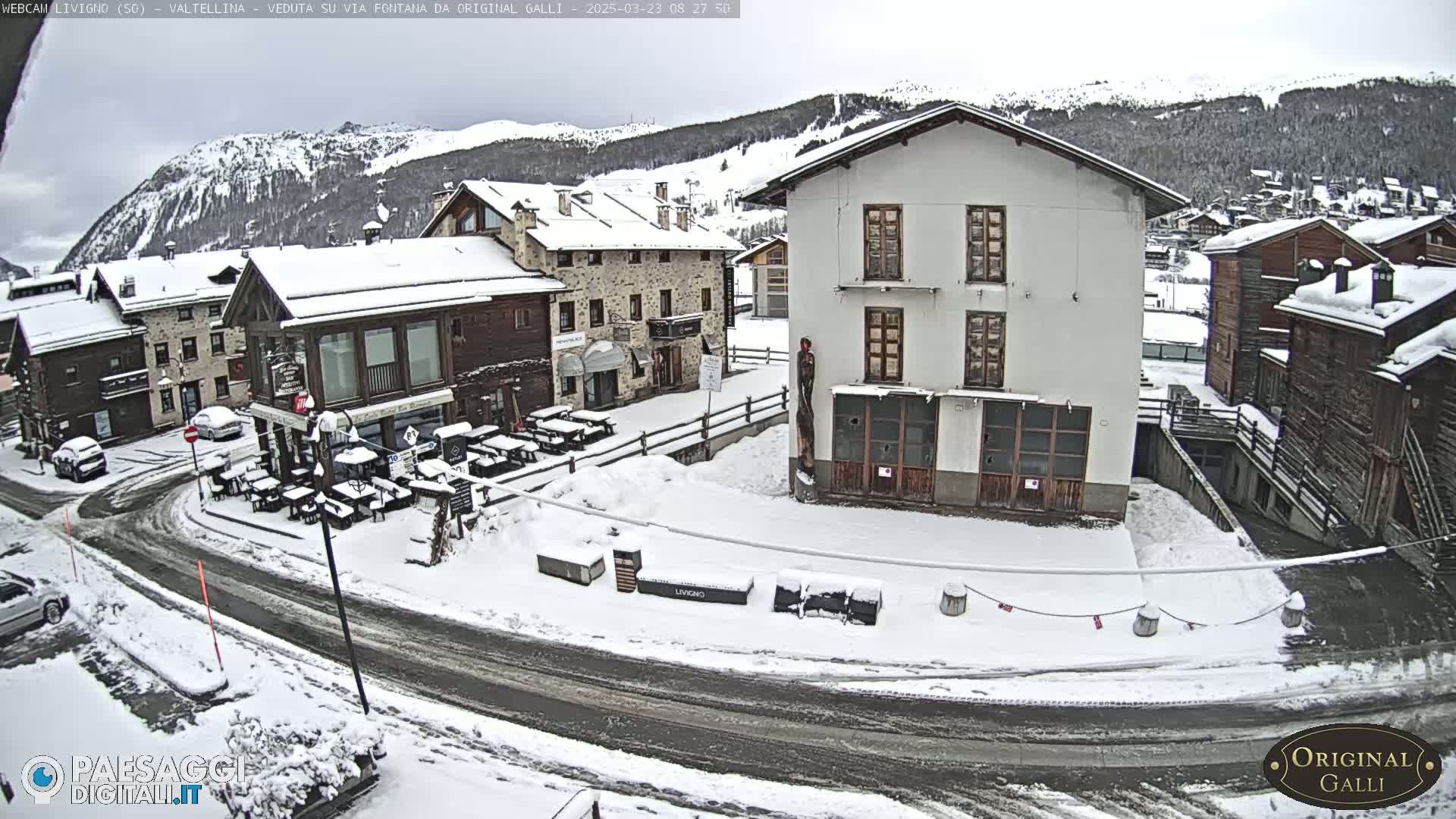 A snow-covered village square with several buildings, some with outdoor seating areas, is seen under an overcast sky, with mountains in the background.