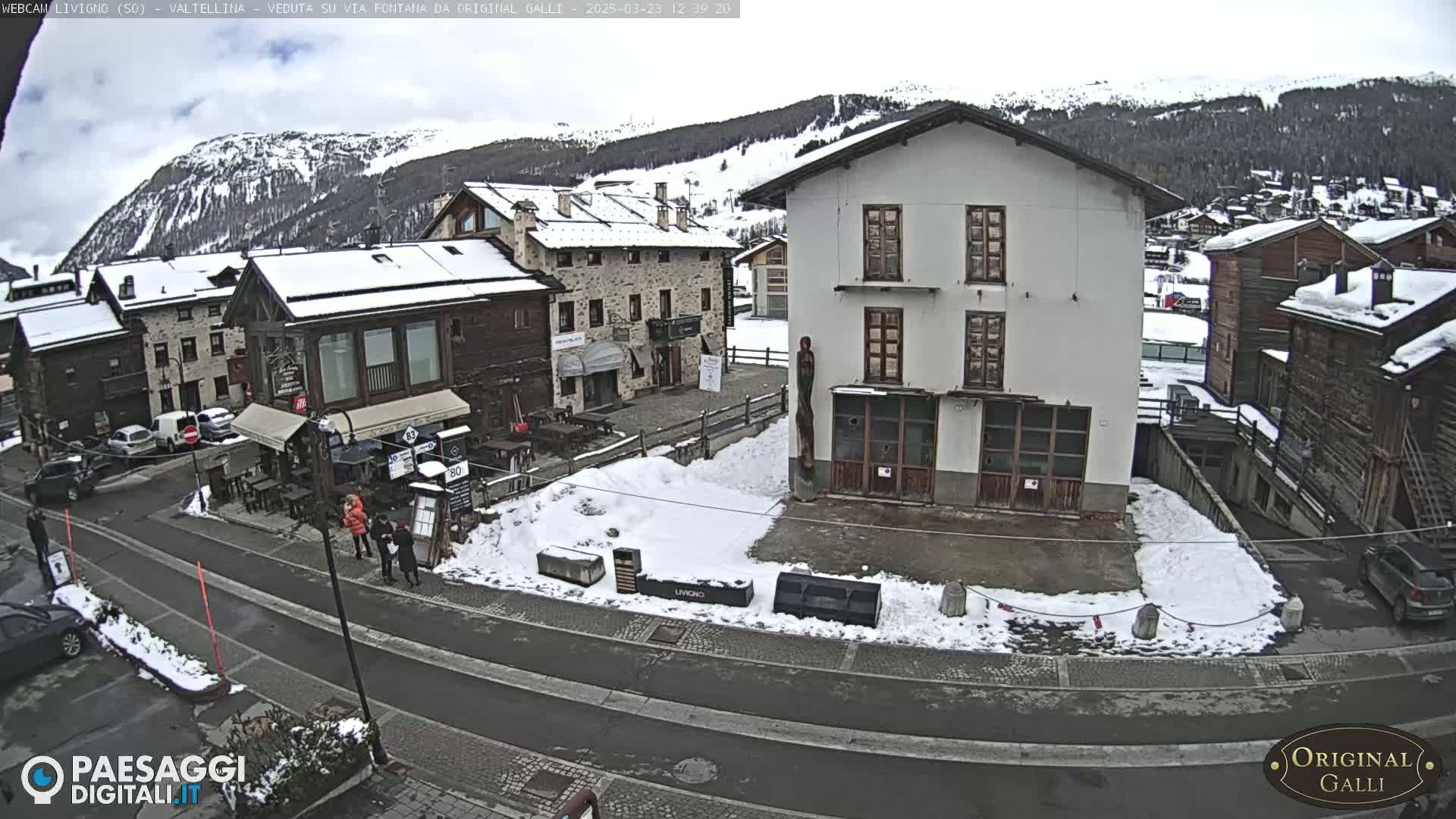 A snow-covered village square is viewed from above, with several buildings, parked cars, and a few pedestrians visible under an overcast sky.