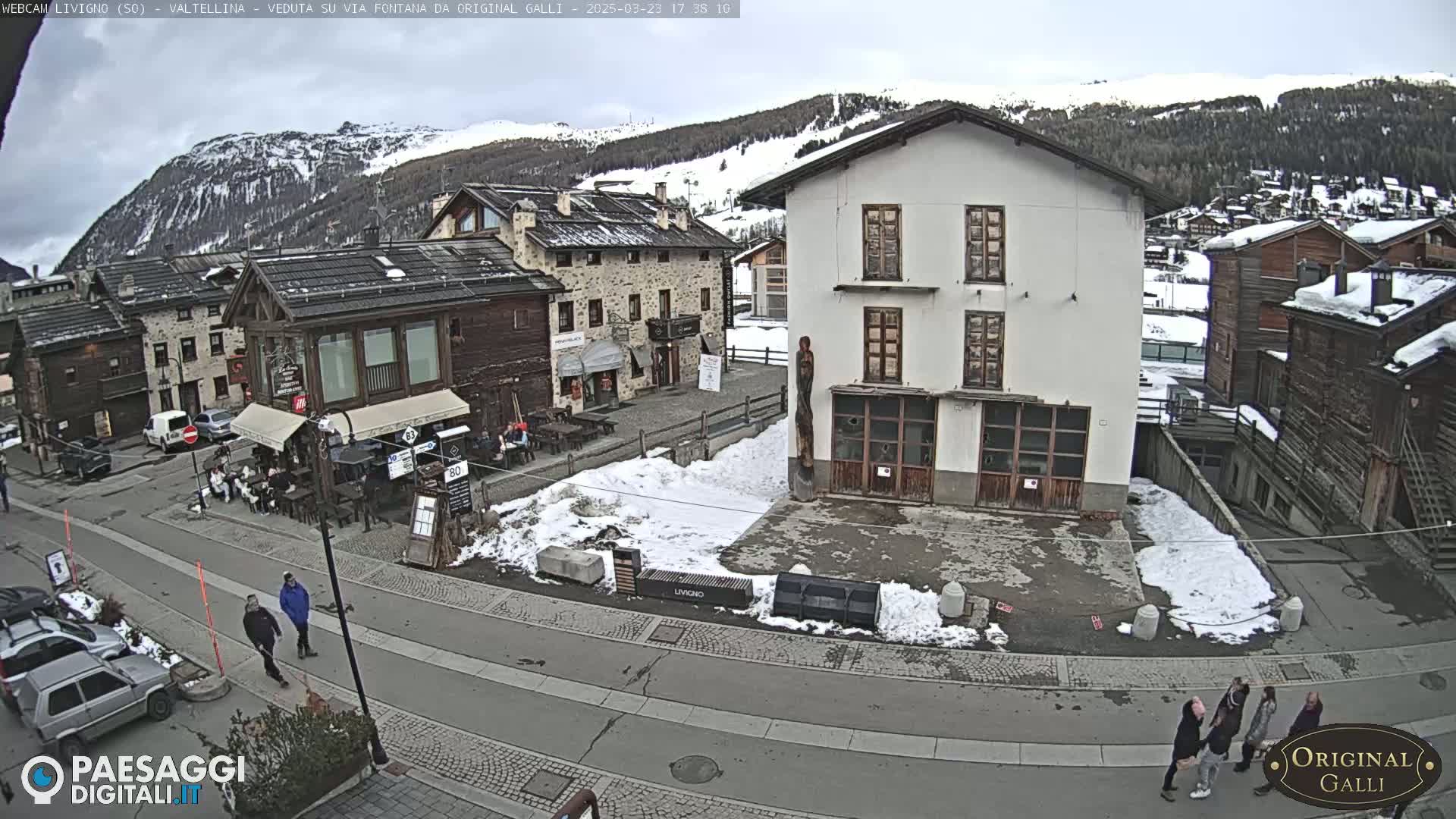 A snow-dusted village square in the mountains, under an overcast sky, features several buildings, some people walking, and parked cars.