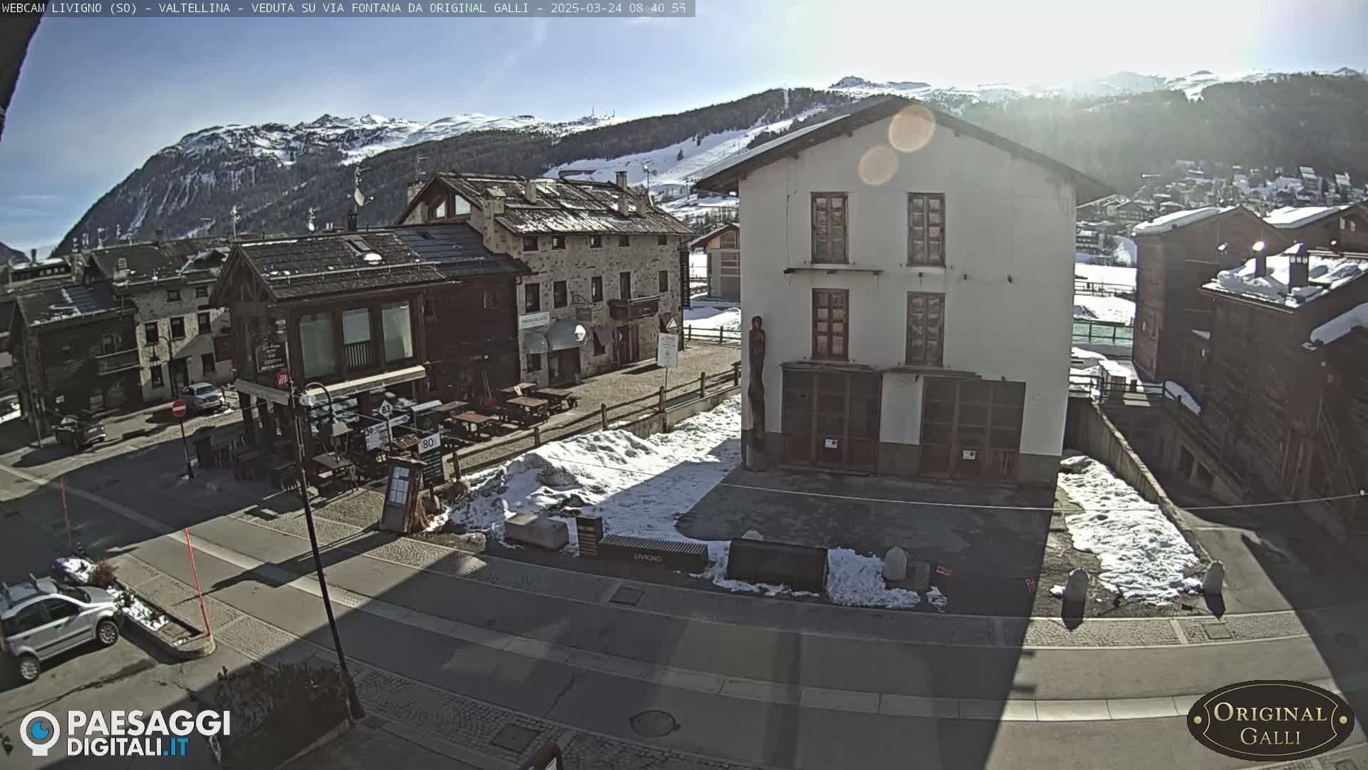 A sunny day in a snow-dusted mountain village shows a town square with buildings, some with snow on the ground.