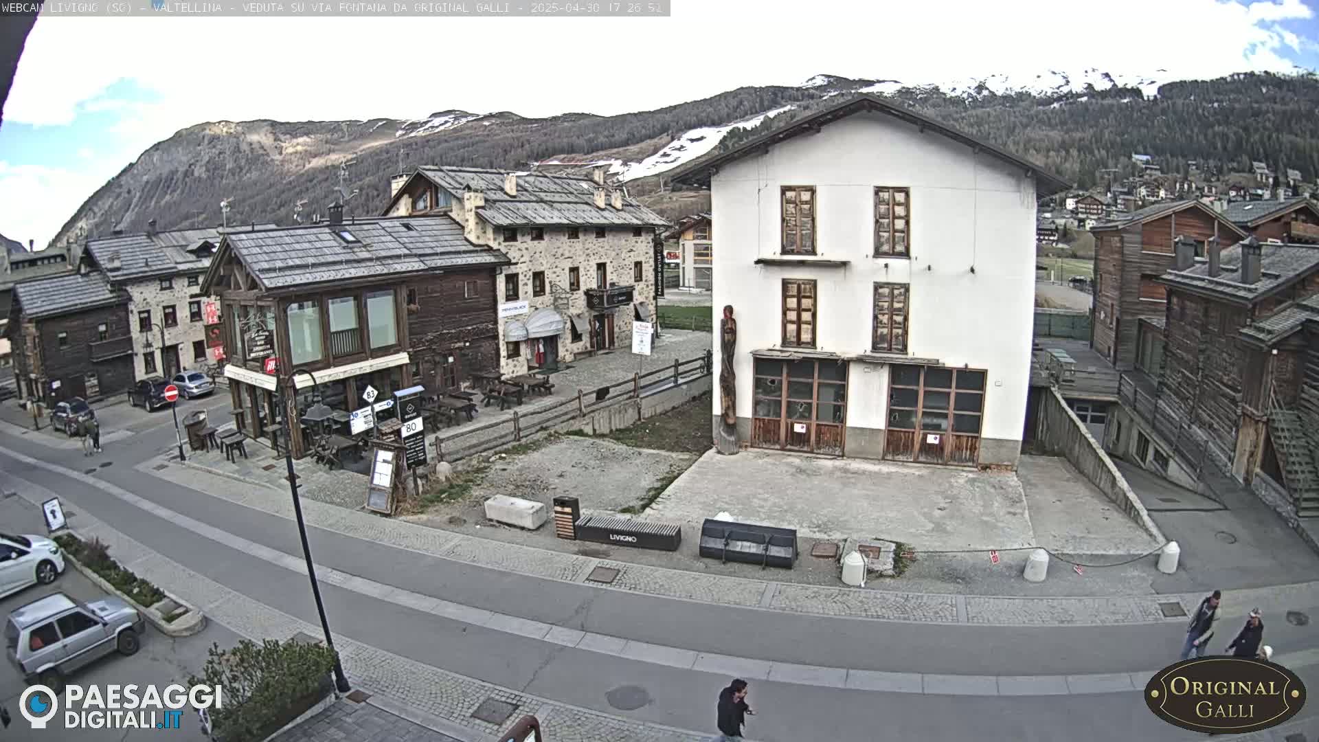 A mostly sunny view of a village square nestled in a valley with snow-capped mountains in the background.