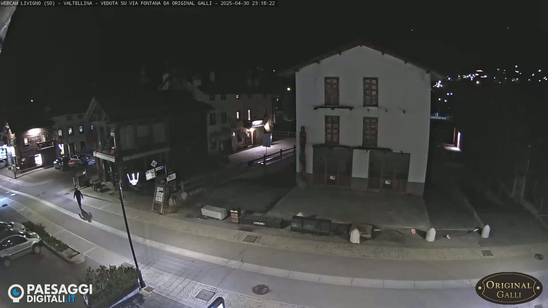 A nighttime, high-angle view of a sparsely populated town square with a few buildings, a person walking on a street, and some parked cars.