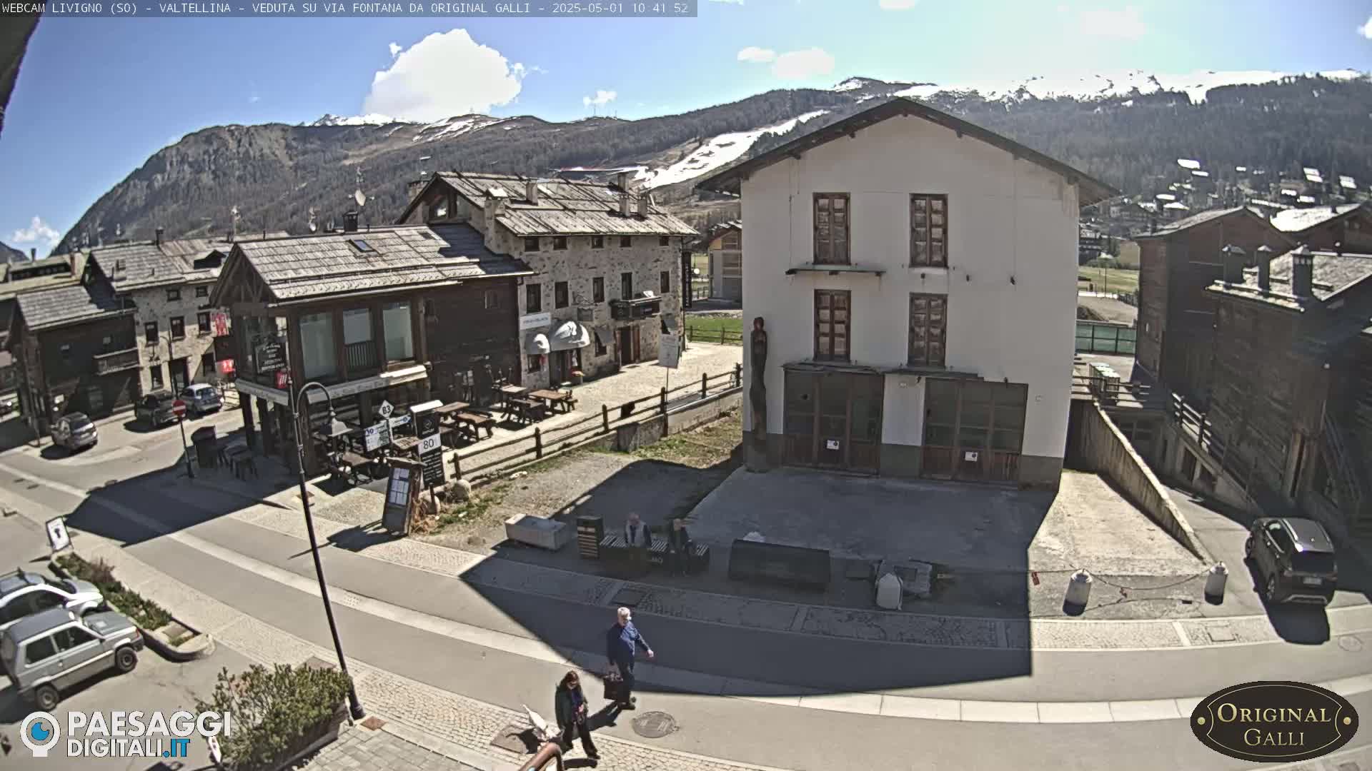 A sunny day in a mountain village, showing stone buildings, a paved road, and a few people walking.