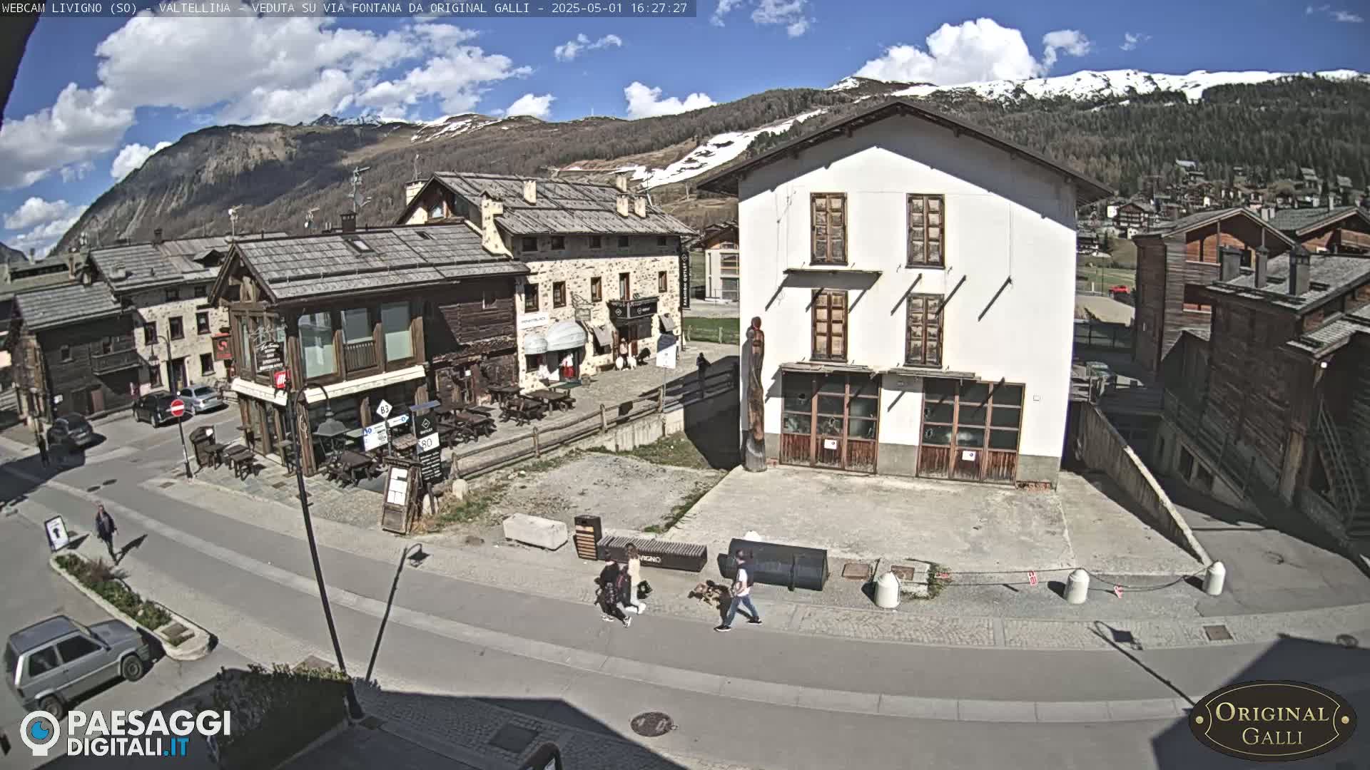 A sunny day in a mountain village square with several buildings, a few people walking, and cars parked.