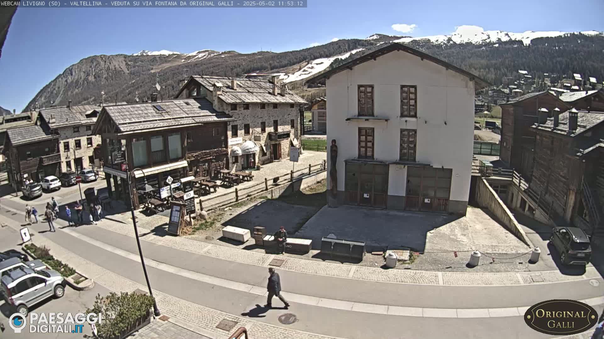 A sunny mountain village scene shows a paved road with parked cars and pedestrians, alongside stone buildings, with snow-capped mountains in the background.