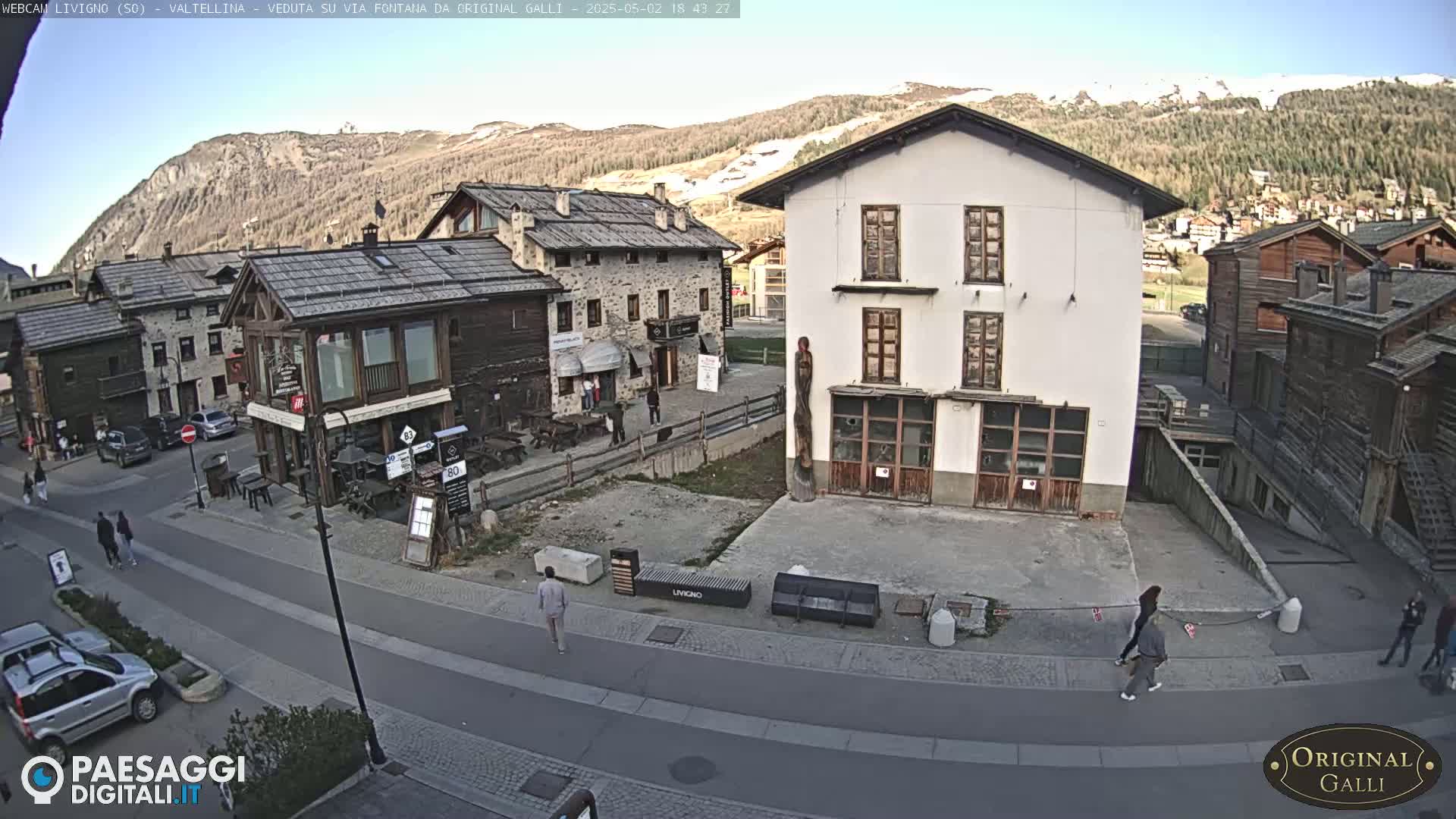 A sunny day in a mountain village shows a street scene with several buildings, some pedestrians, and cars parked along the side of the road.
