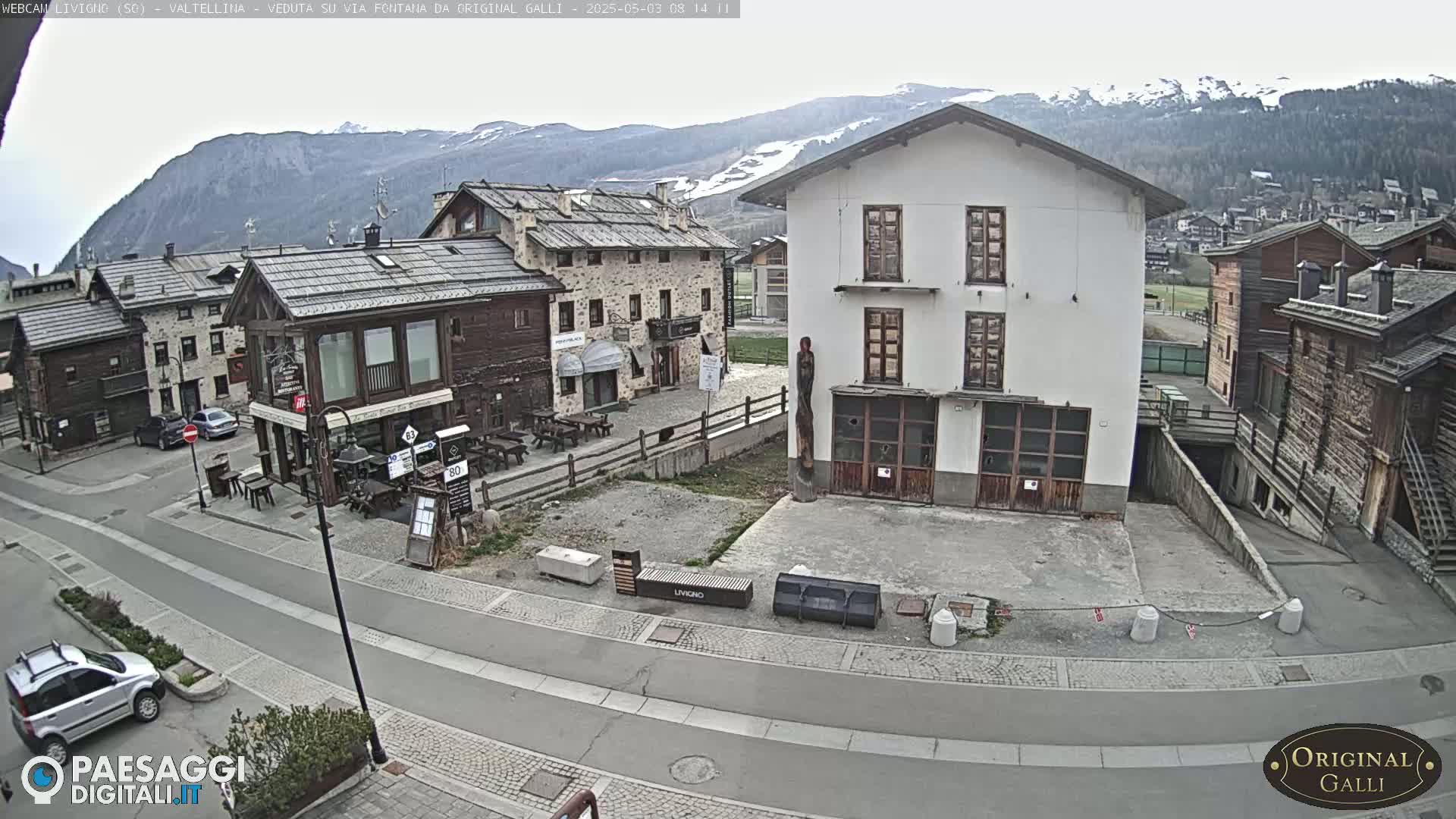 A mostly overcast day reveals a village square with several buildings, a few cars, and mountains in the background.